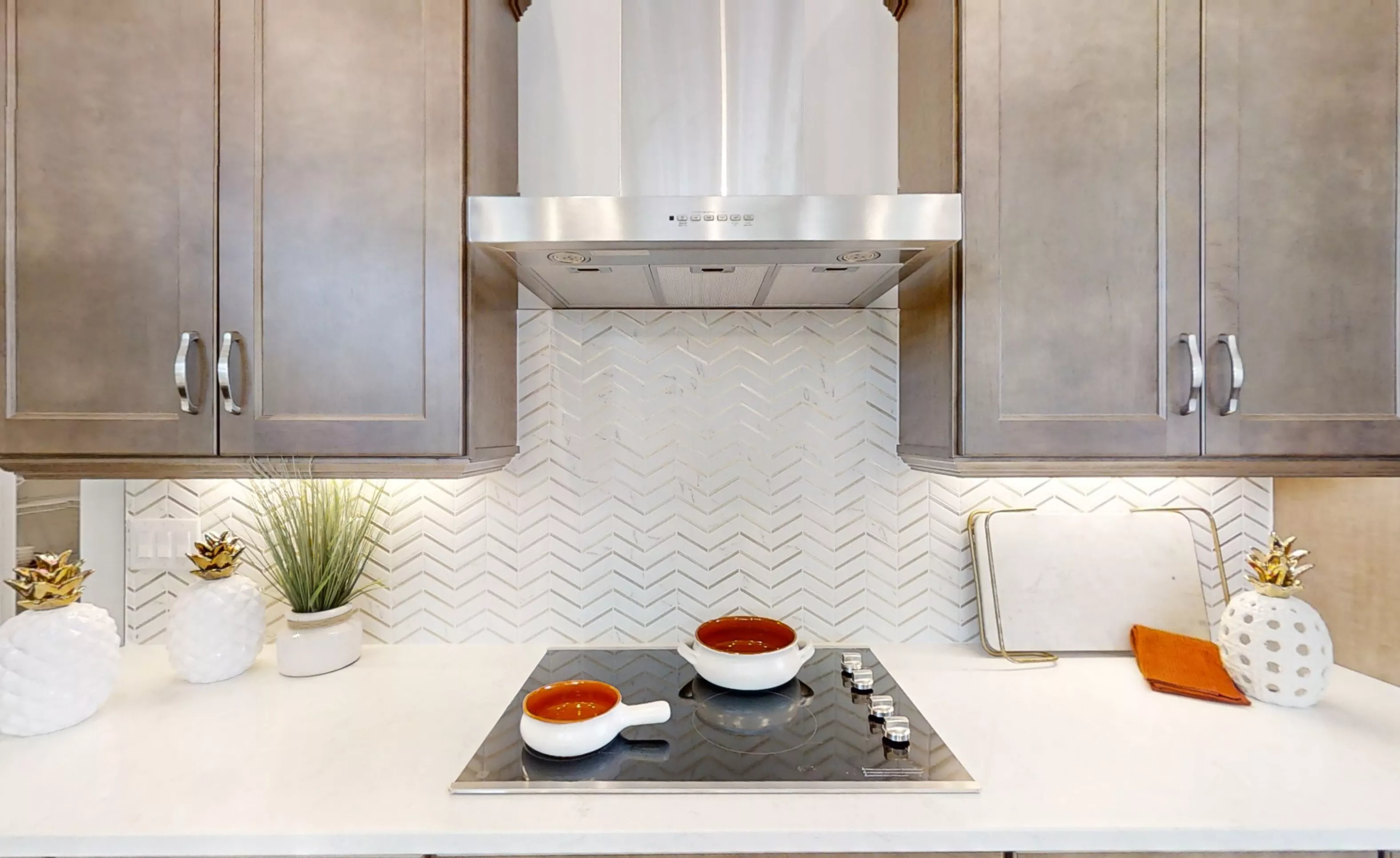Kitchen stovetop with stainless steel range hood, grey cabinets, white countertops, and white herringbone backsplash.