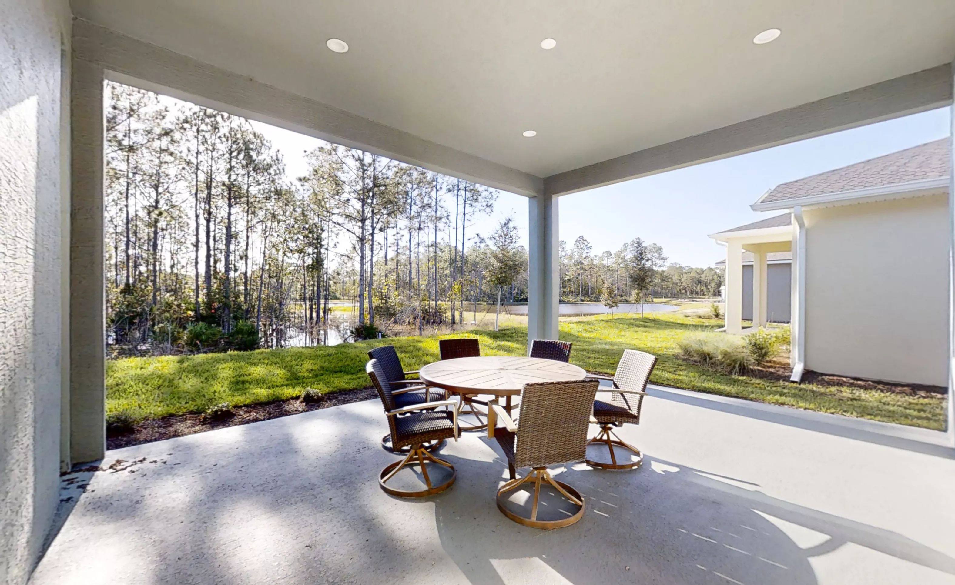 Covered concrete patio with a round dining table and chairs, overlooking a grassy yard, trees, and a small pond.