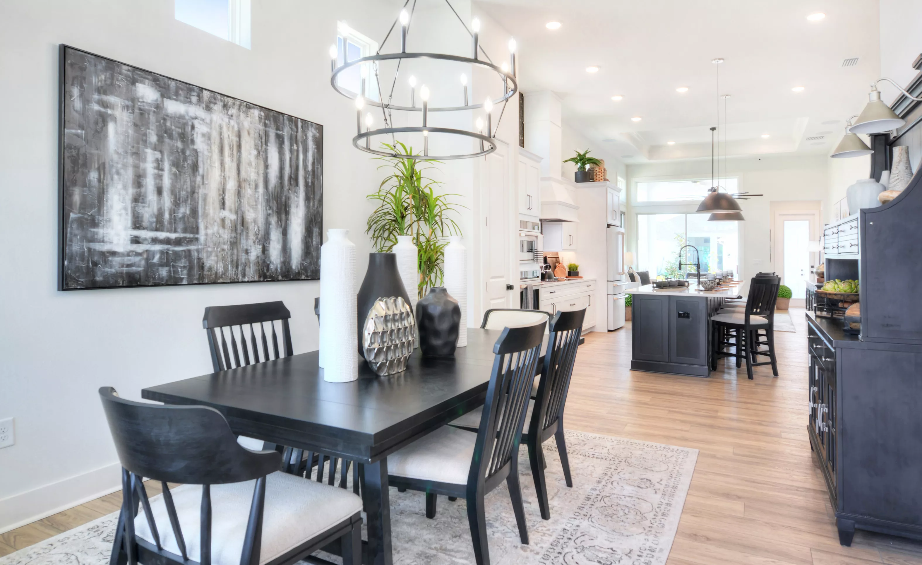 Dining room of the Lantana plan with a black table, spindle chairs, and a large metal chandelier, opening to a kitchen.