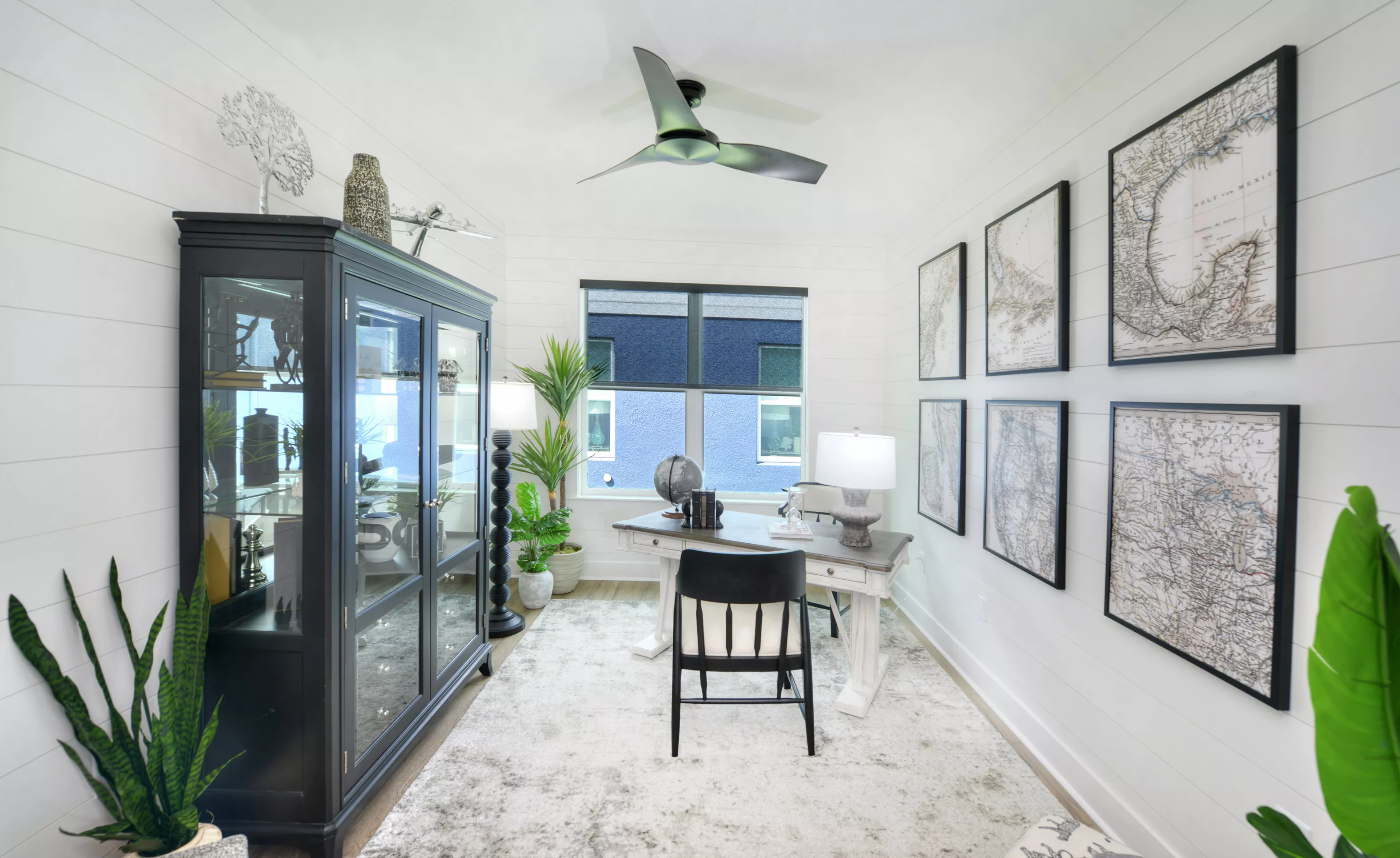 Lantana home office with white shiplap walls, black display cabinet, wood desk, and framed map wall art.