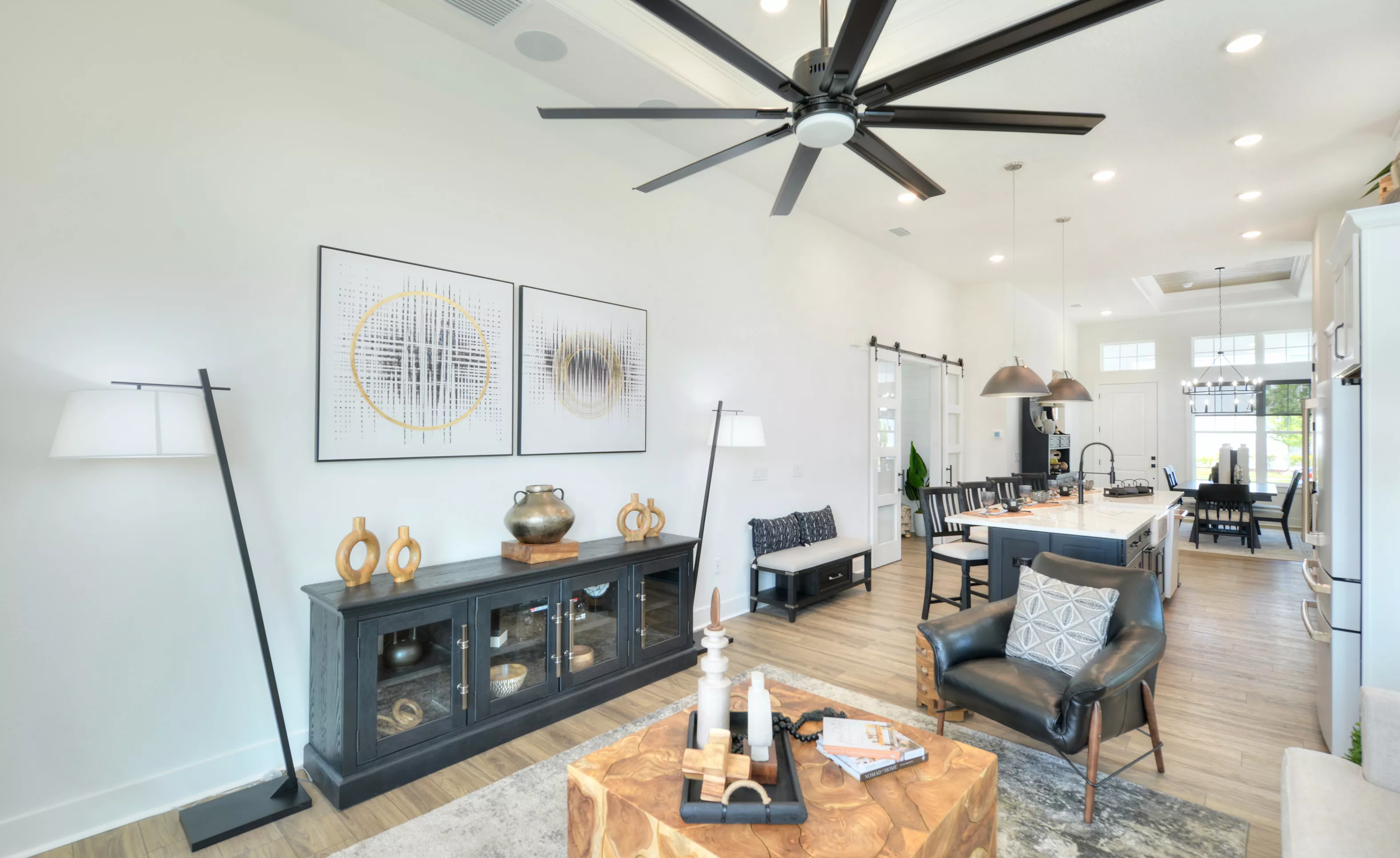 Living room of the Lantana model featuring a large black ceiling fan, wood-look flooring, and an open-concept kitchen view.