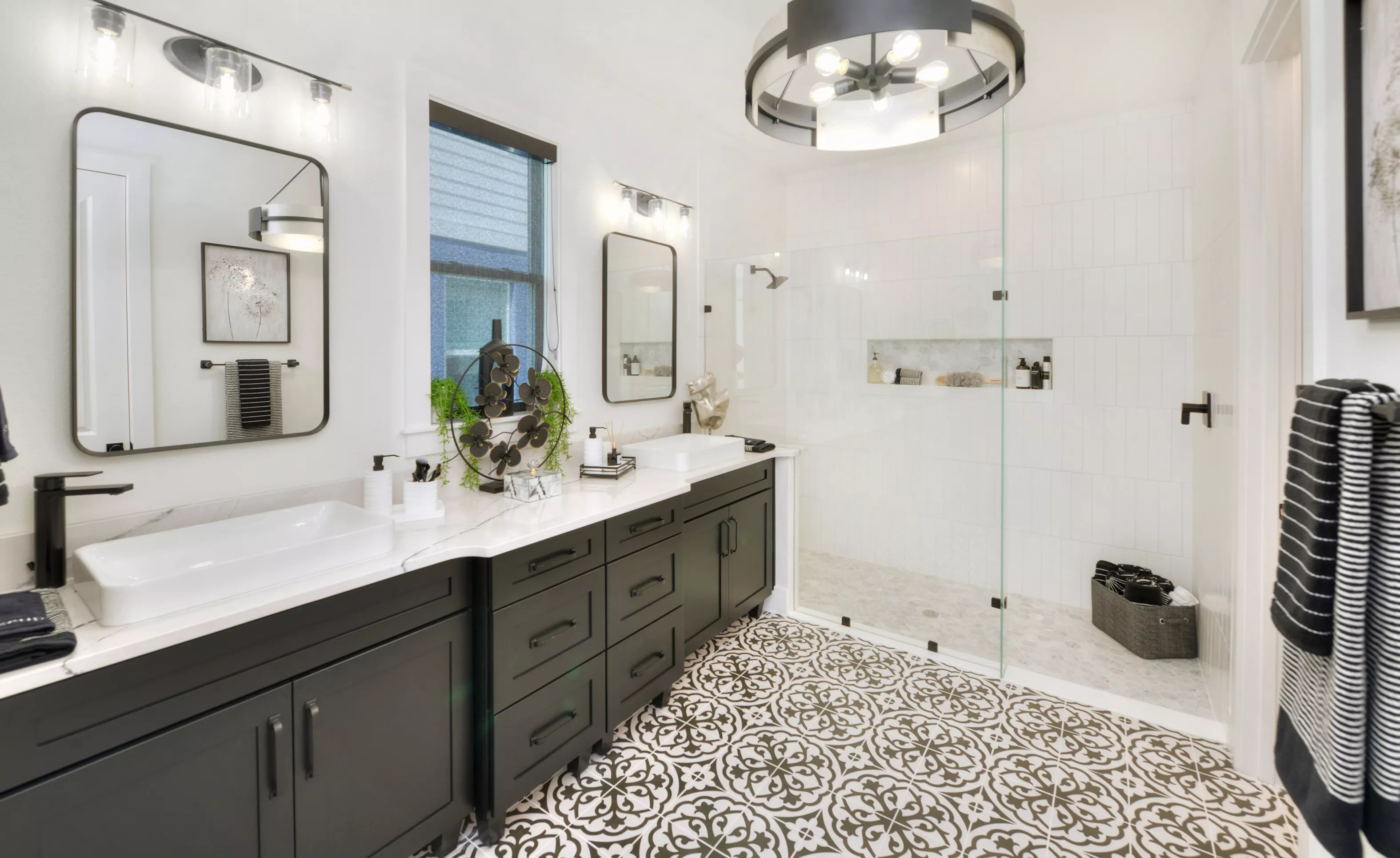 Primary bath with black vanity, white countertops, double vessel sinks, patterned tile floors, and a glass-enclosed shower.
