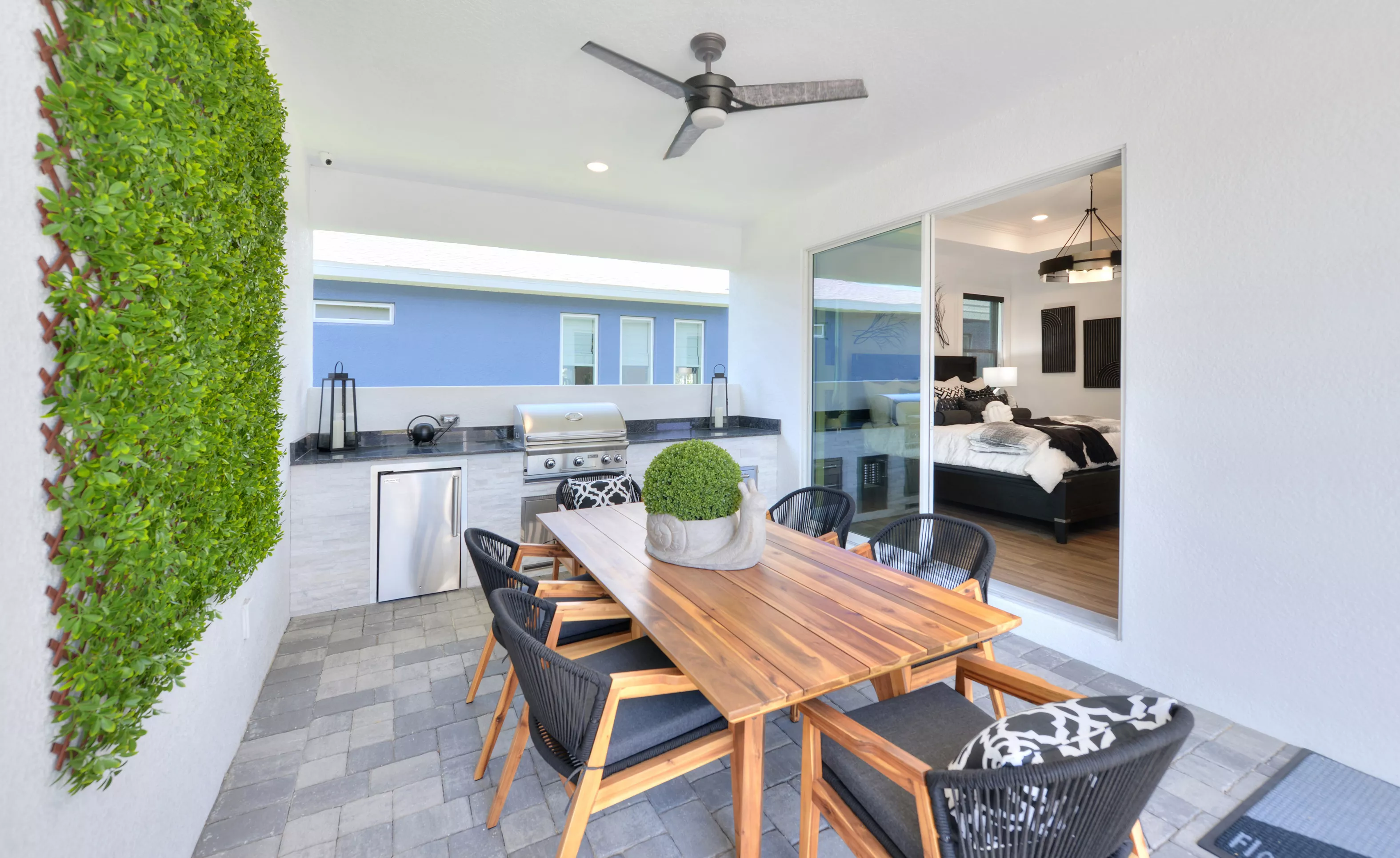 Covered outdoor patio with a dining set, built-in grill area, green wall feature, and sliding glass doors to a bedroom.
