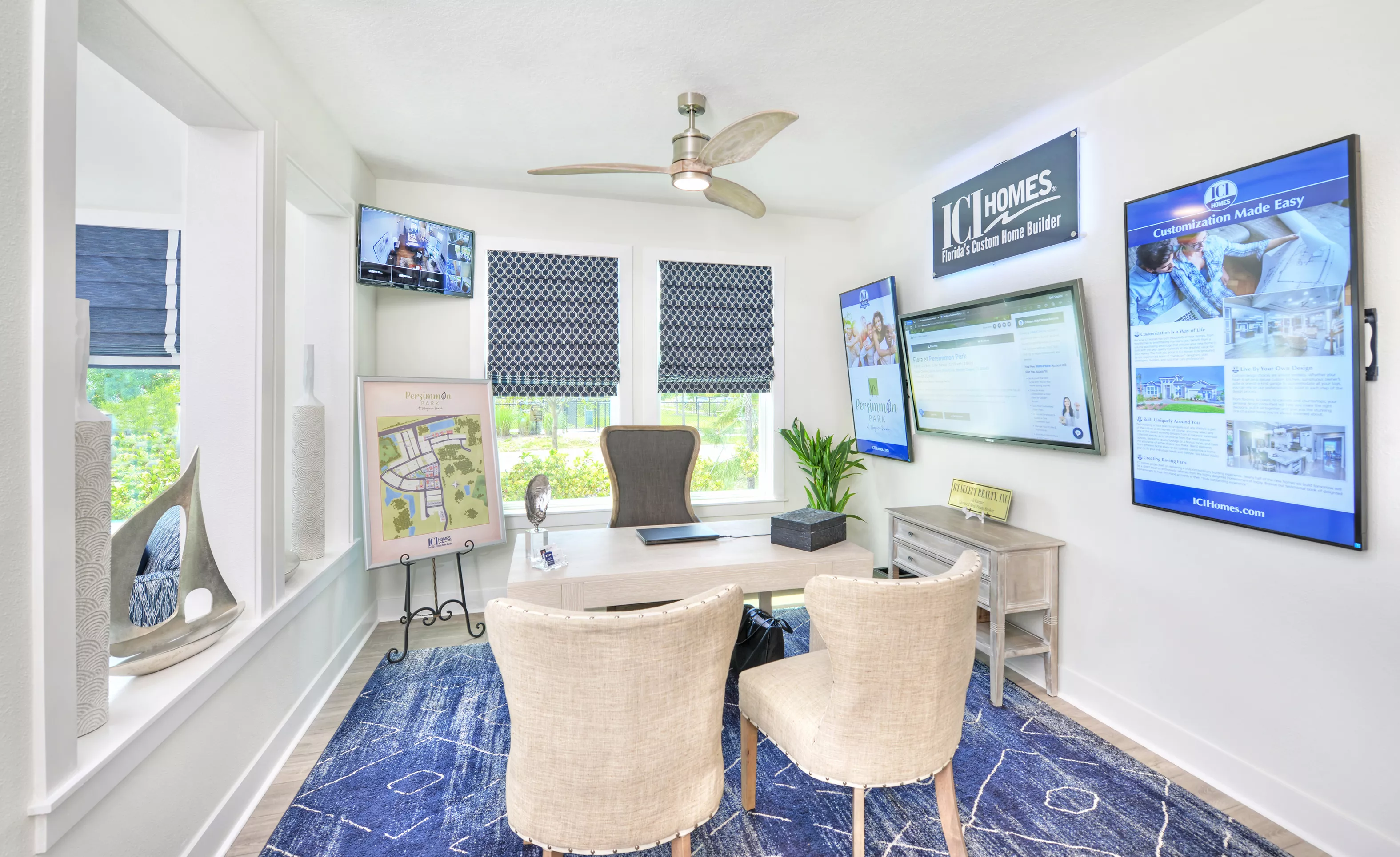 Azalea home office at Persimmon Park with a light wood desk, two tufted chairs, patterned blue rug, and wall-mounted screens.