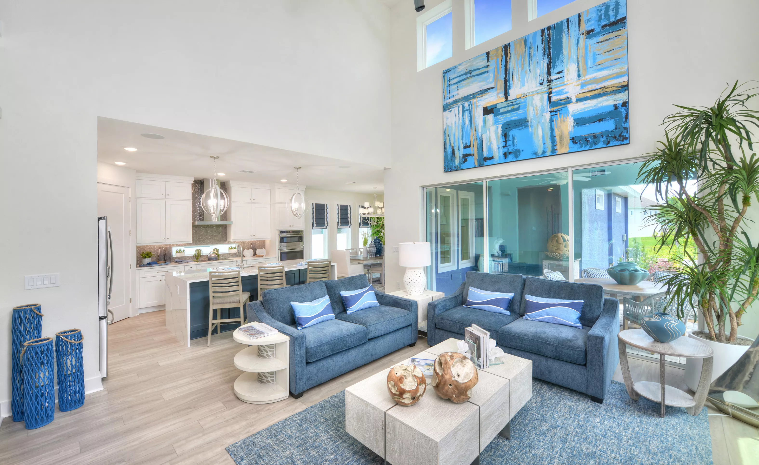 Living room in Azalea floor plan with vaulted ceilings, large blue wall art, blue sofas, and view into the open kitchen.