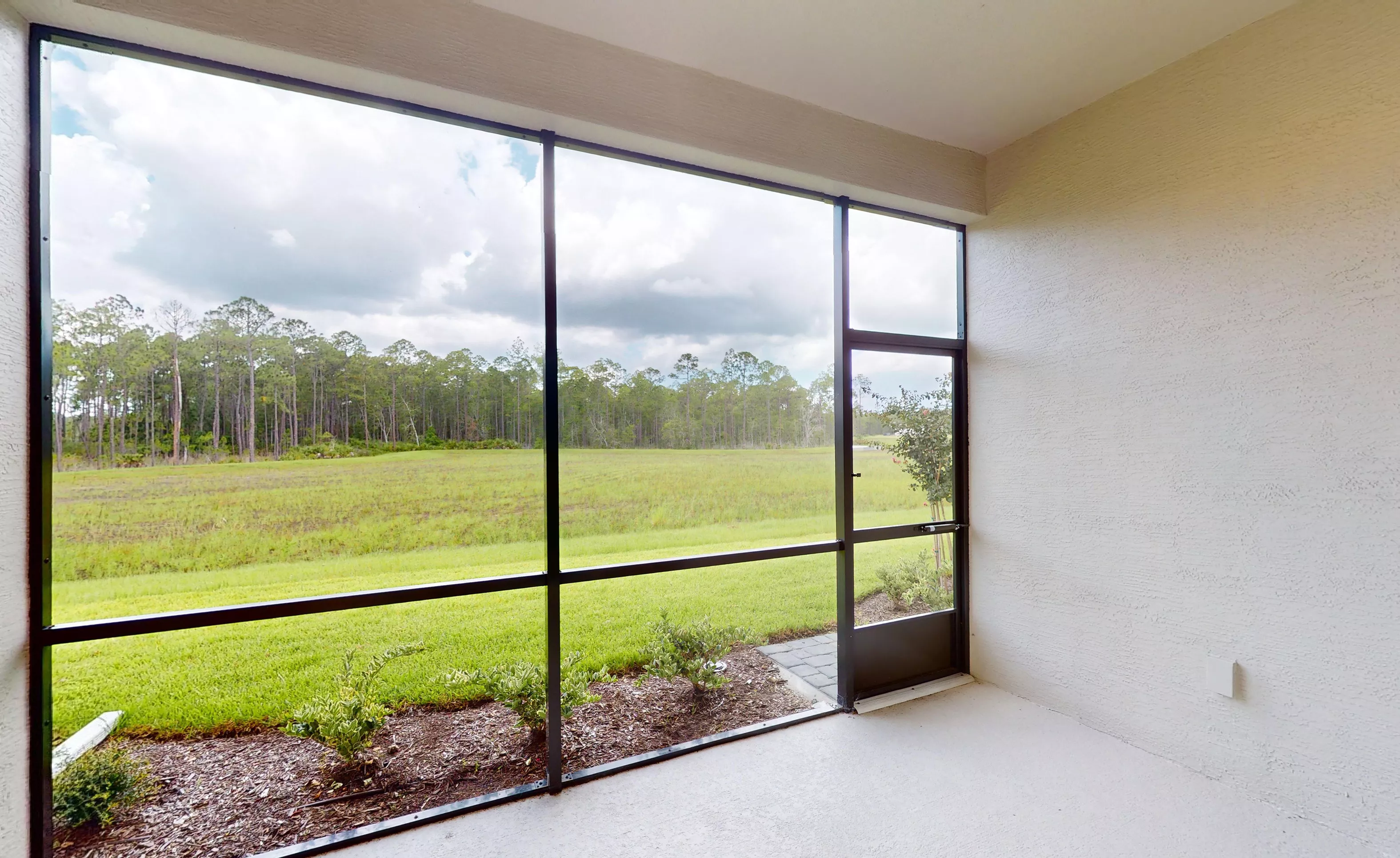 Screened lanai with a concrete floor and a view of a grassy lawn leading to a wooded area.