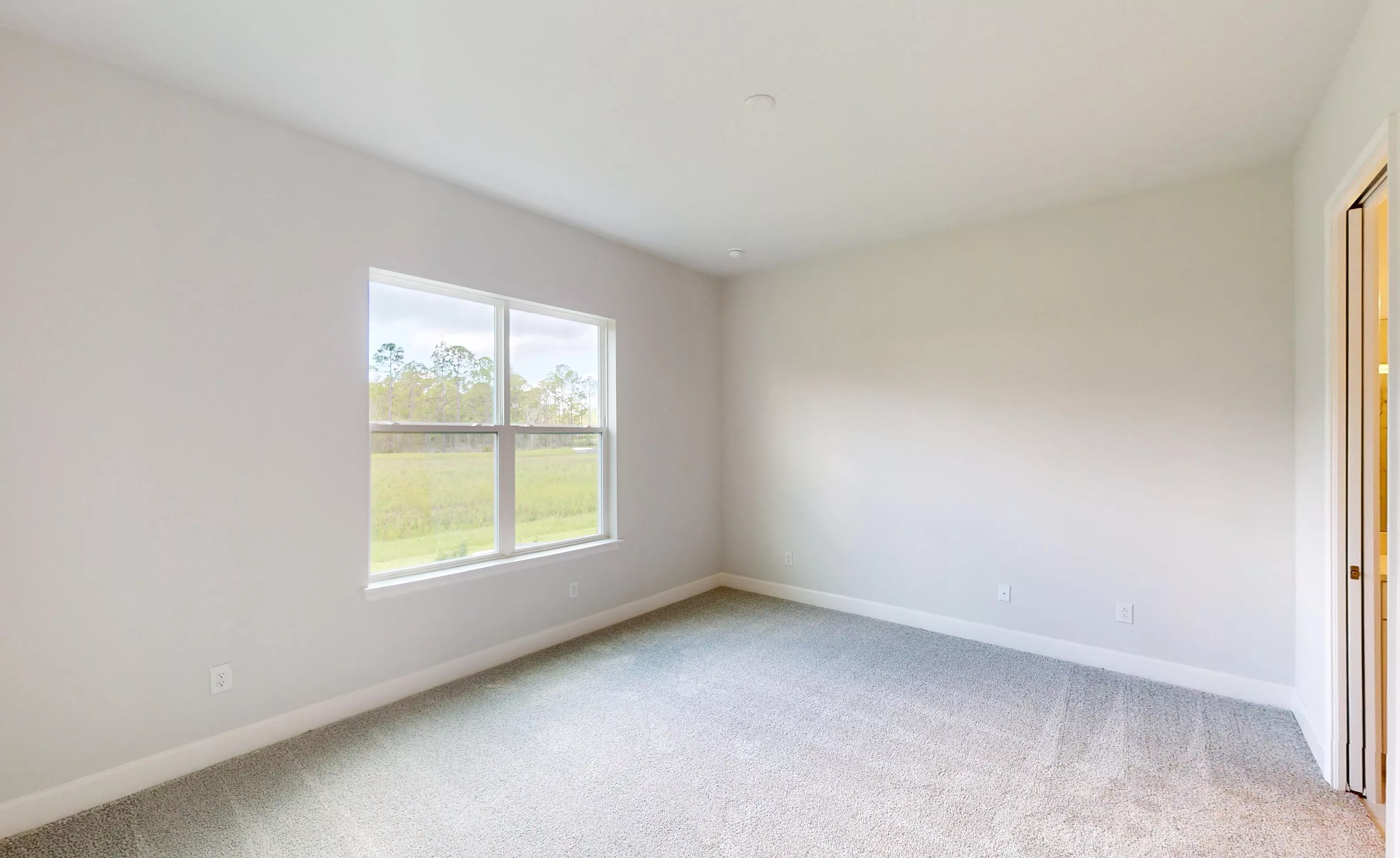 Empty secondary bedroom in the Blossom II plan with light gray walls, carpeted floors, and a window overlooking greenery.