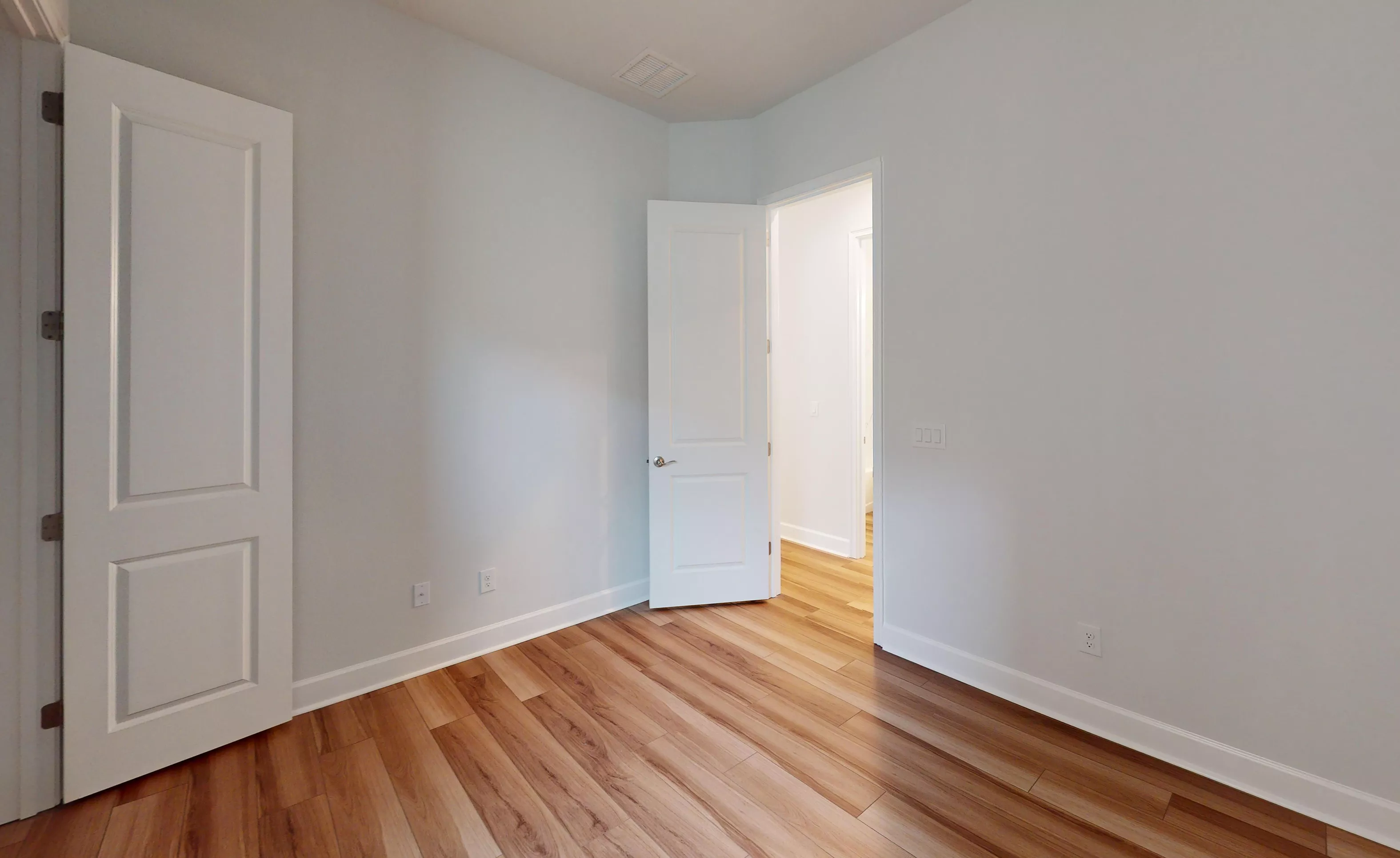 Empty secondary bedroom with light gray walls, white trim, and wood-look flooring, showing an open doorway to a hallway.