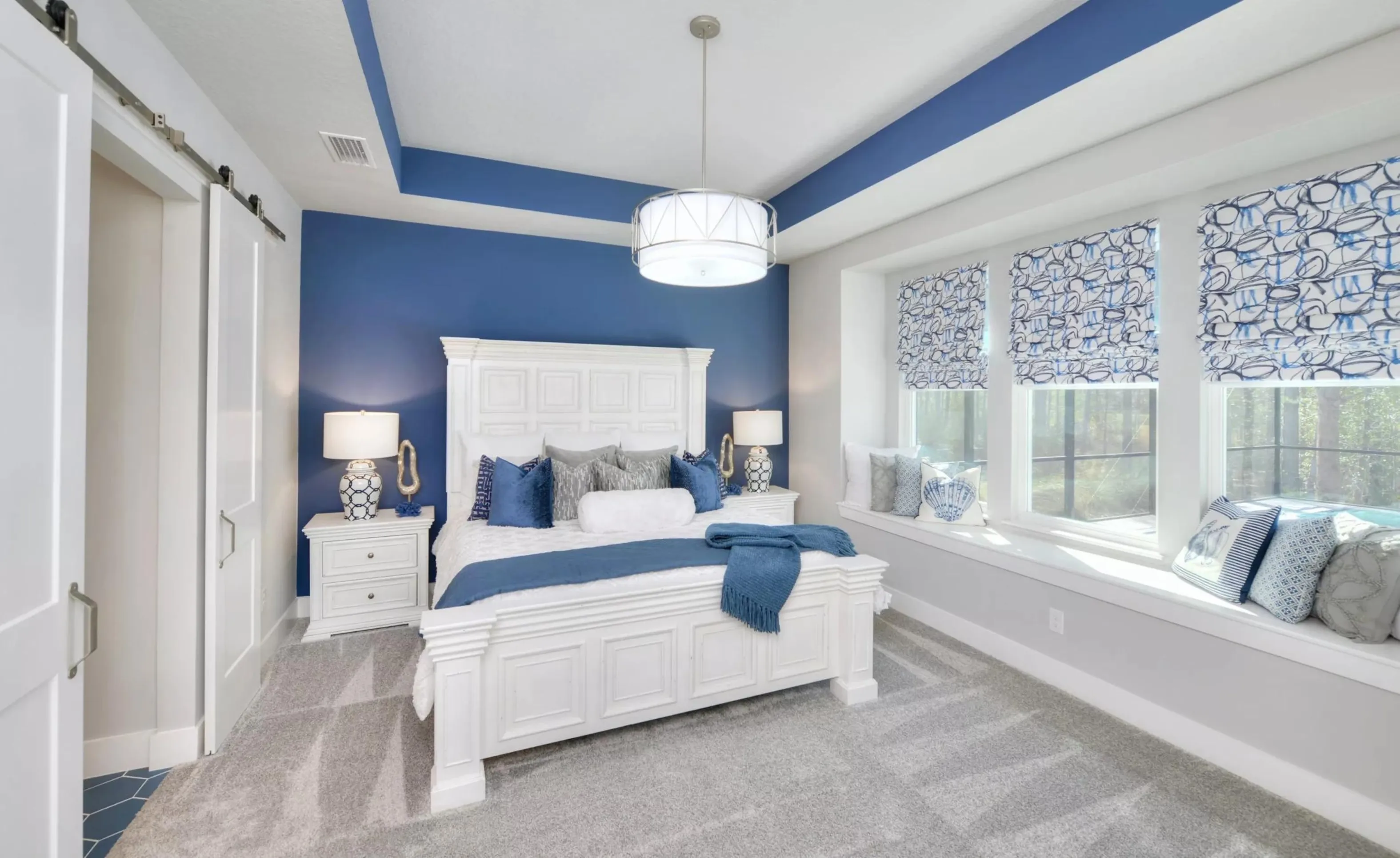 Bedroom in Woodhaven with a white bed, navy blue accent wall, tray ceiling, and a window seat with patterned shades.