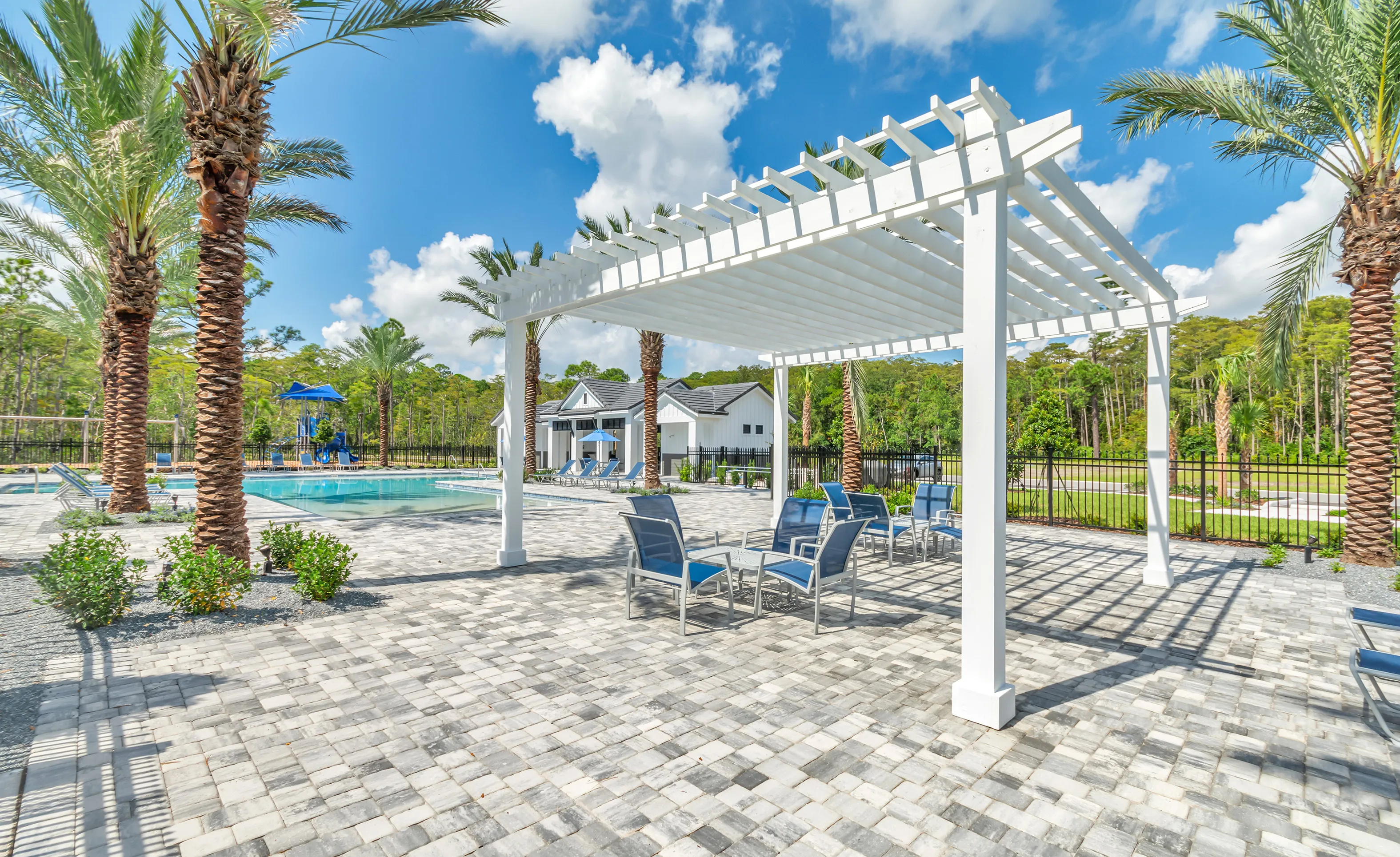 Woodhaven community pool deck with blue chairs under a white pergola, surrounded by palm trees and a clubhouse.