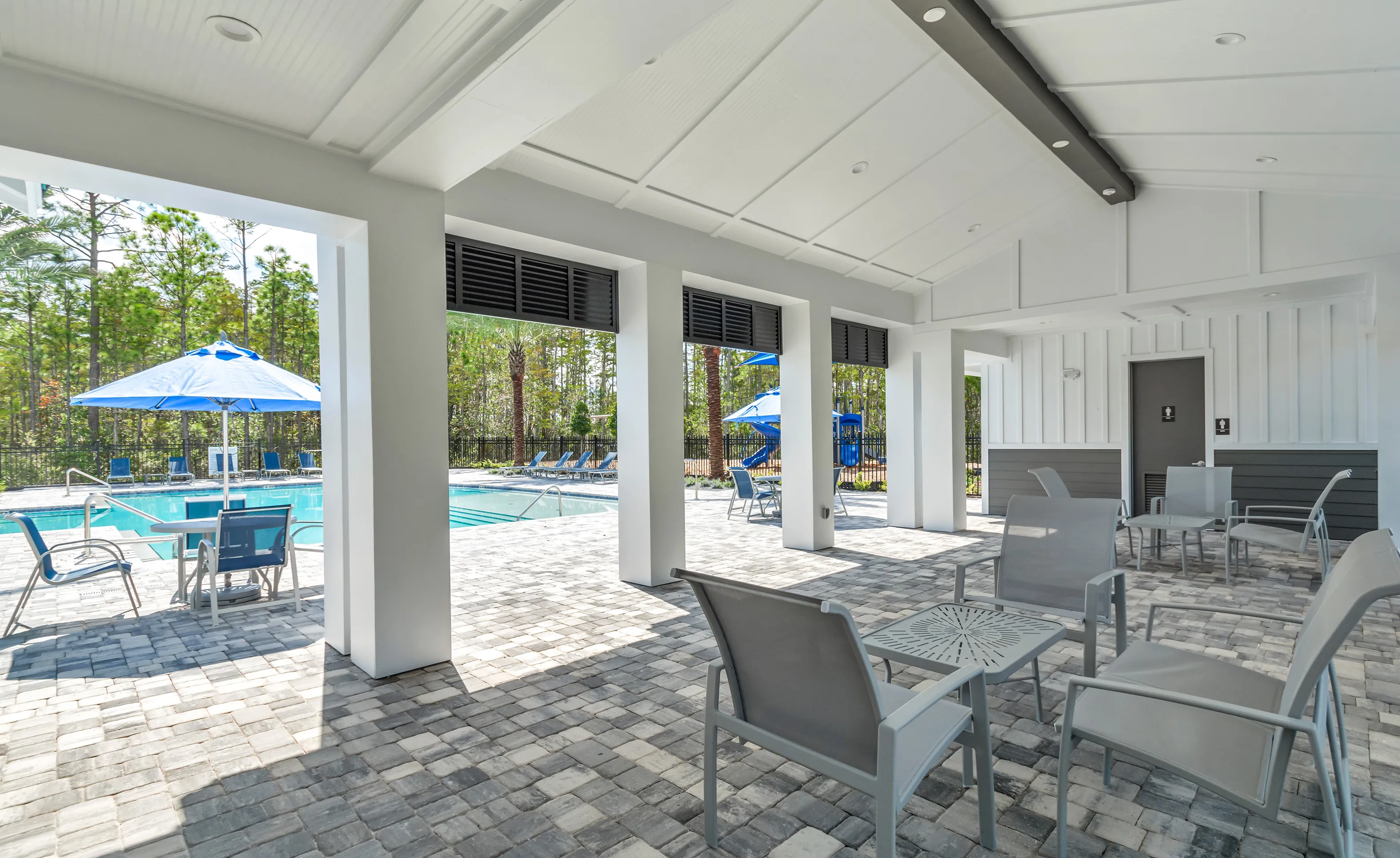 Covered patio at Woodhaven community featuring gray outdoor furniture, paver flooring, and a view of the swimming pool.