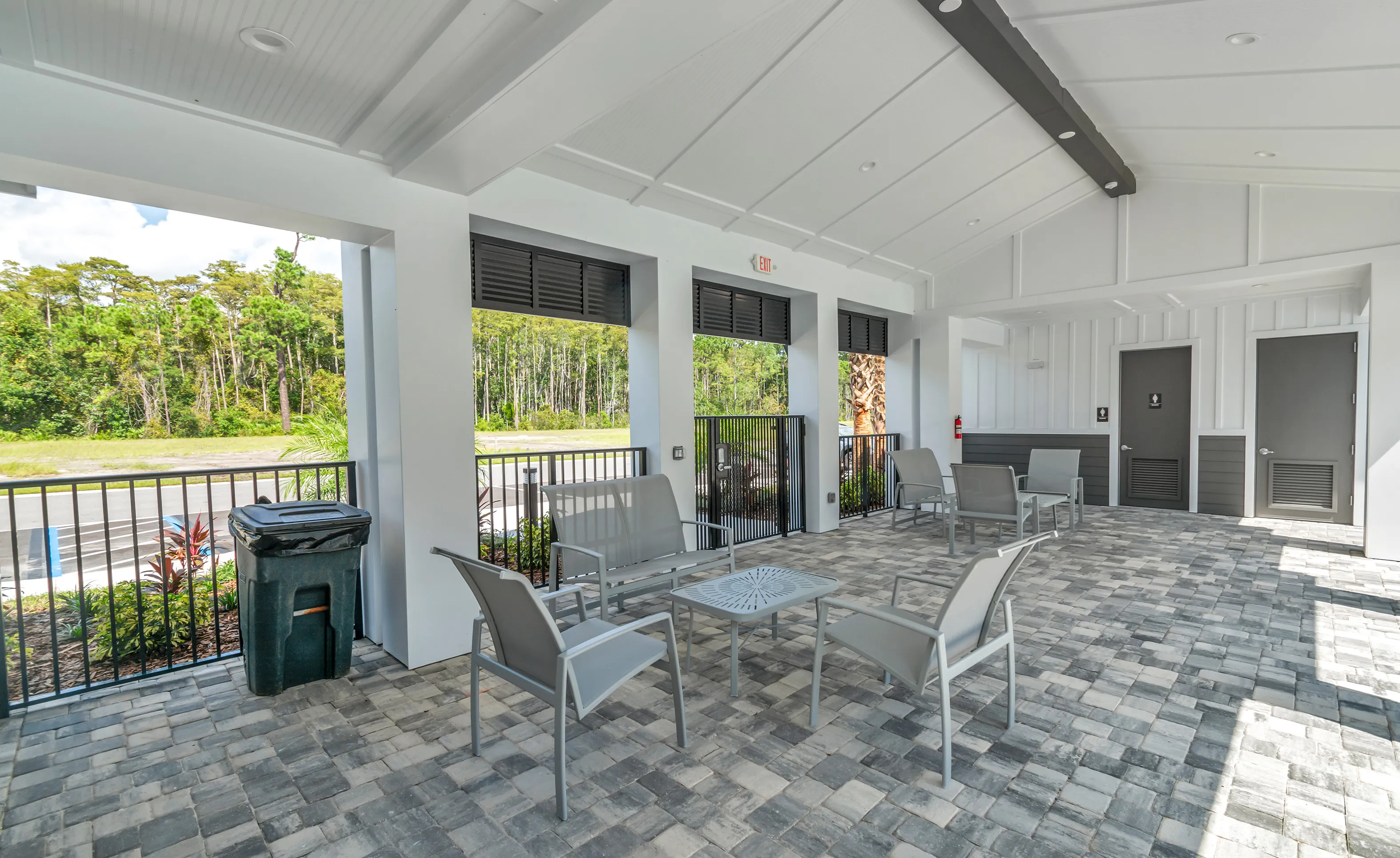 Covered outdoor patio area at Woodhaven with gray metal furniture, paver flooring, and view of trees.