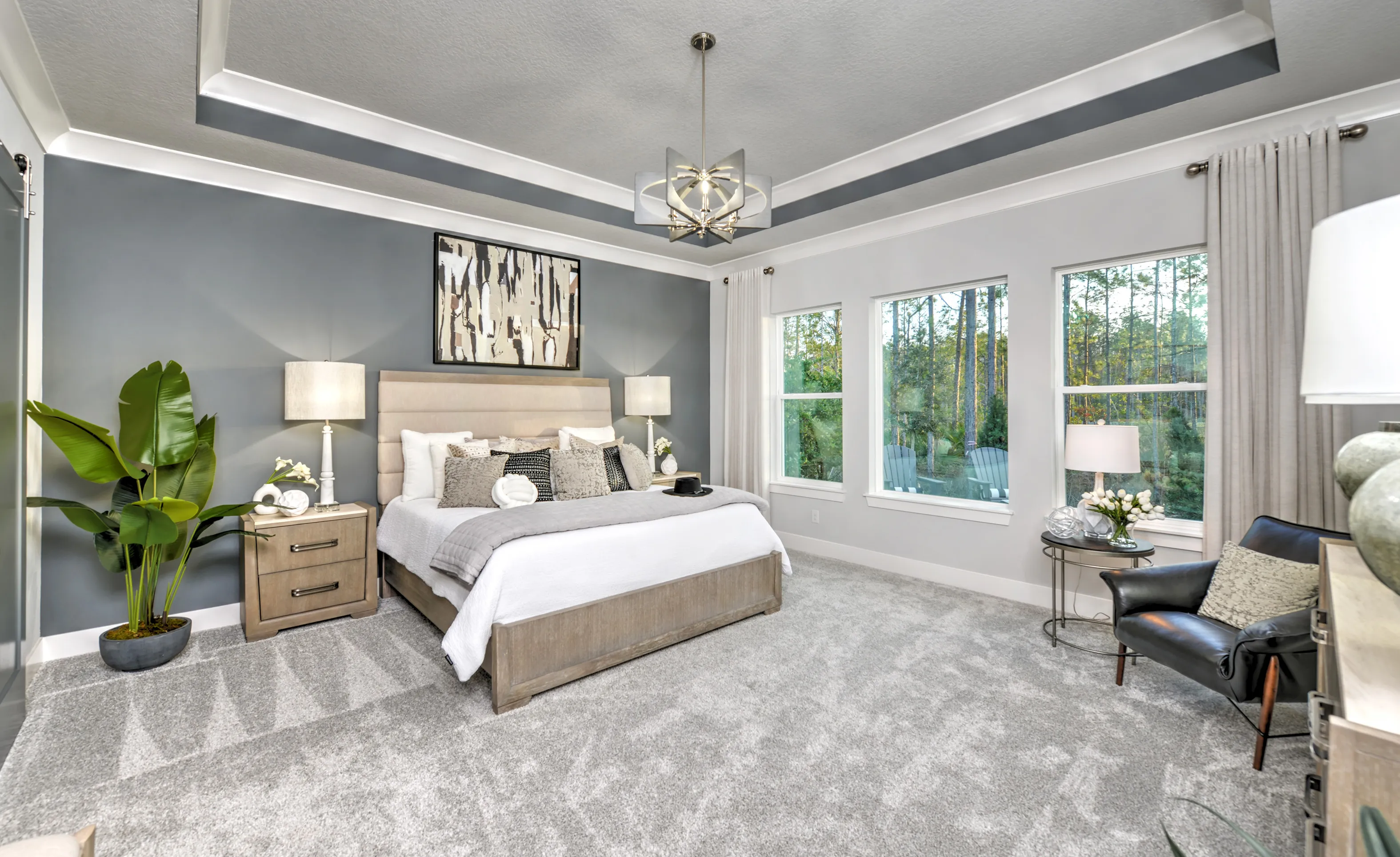 Woodhaven Single Family bedroom with gray tray ceiling, upholstered bed, carpeted floors, and windows overlooking woods.