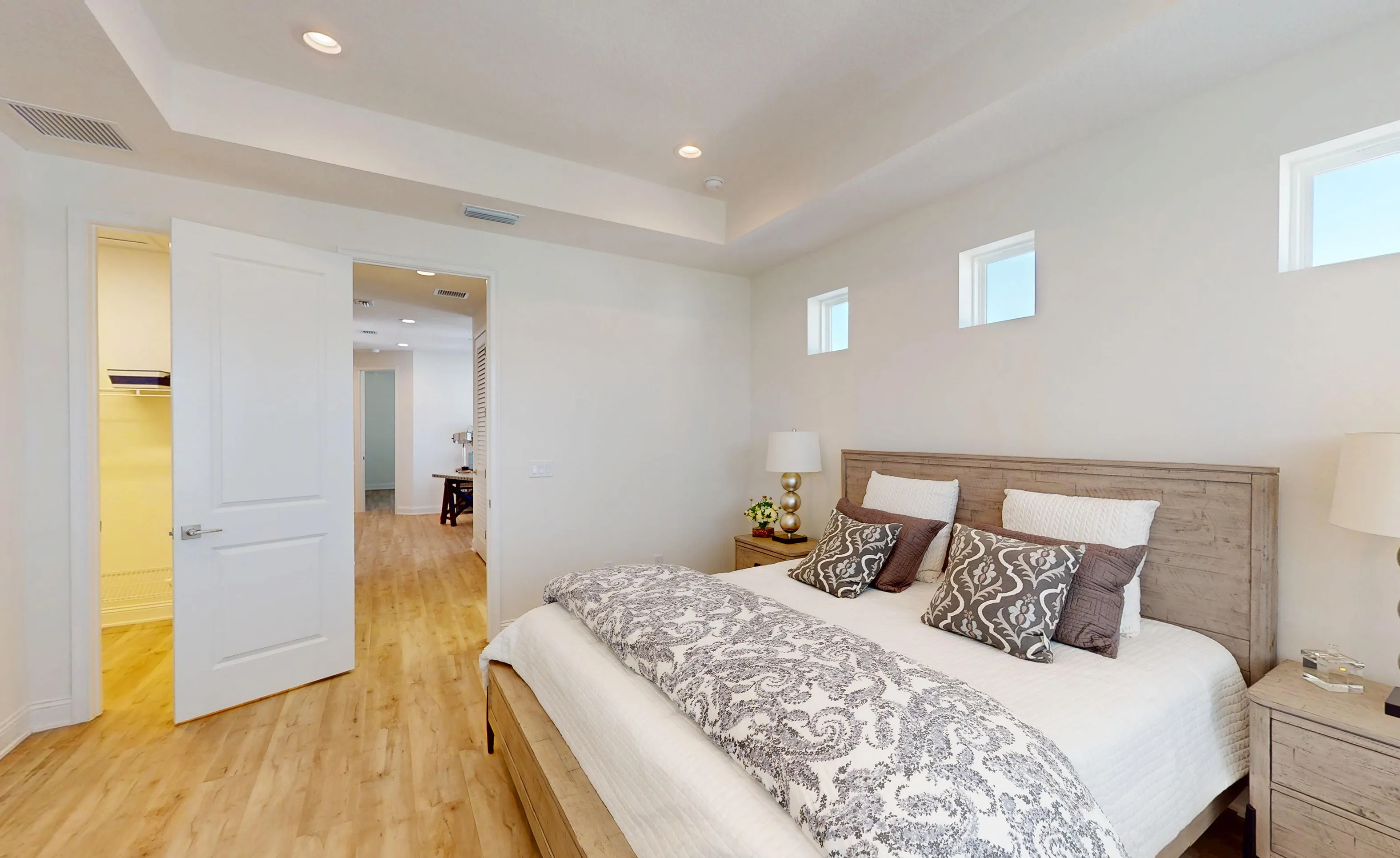 Primary bedroom with wood-toned headboard, tray ceiling, and small square windows above a bed with patterned bedding.
