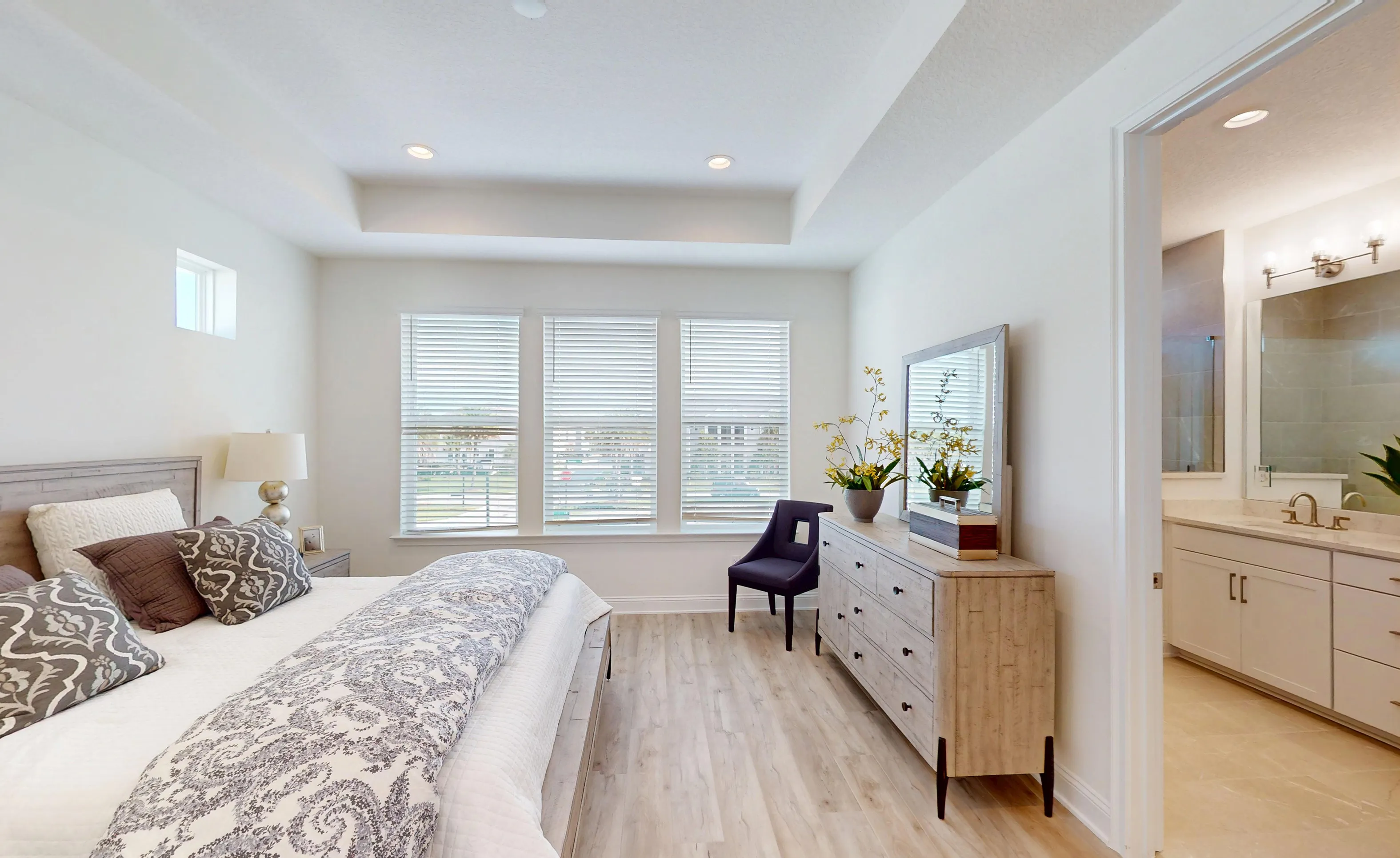 Primary bedroom in Bartram model with tray ceiling, wood-look flooring, three windows with blinds, and attached bathroom.