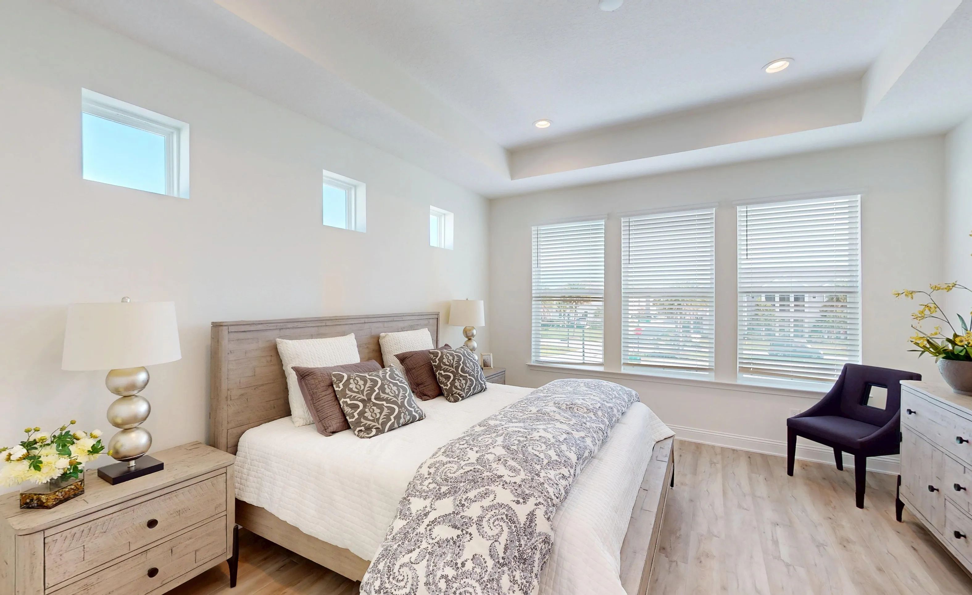 Primary bedroom with a tray ceiling, wood-look flooring, three tall windows with blinds, and small transom windows.