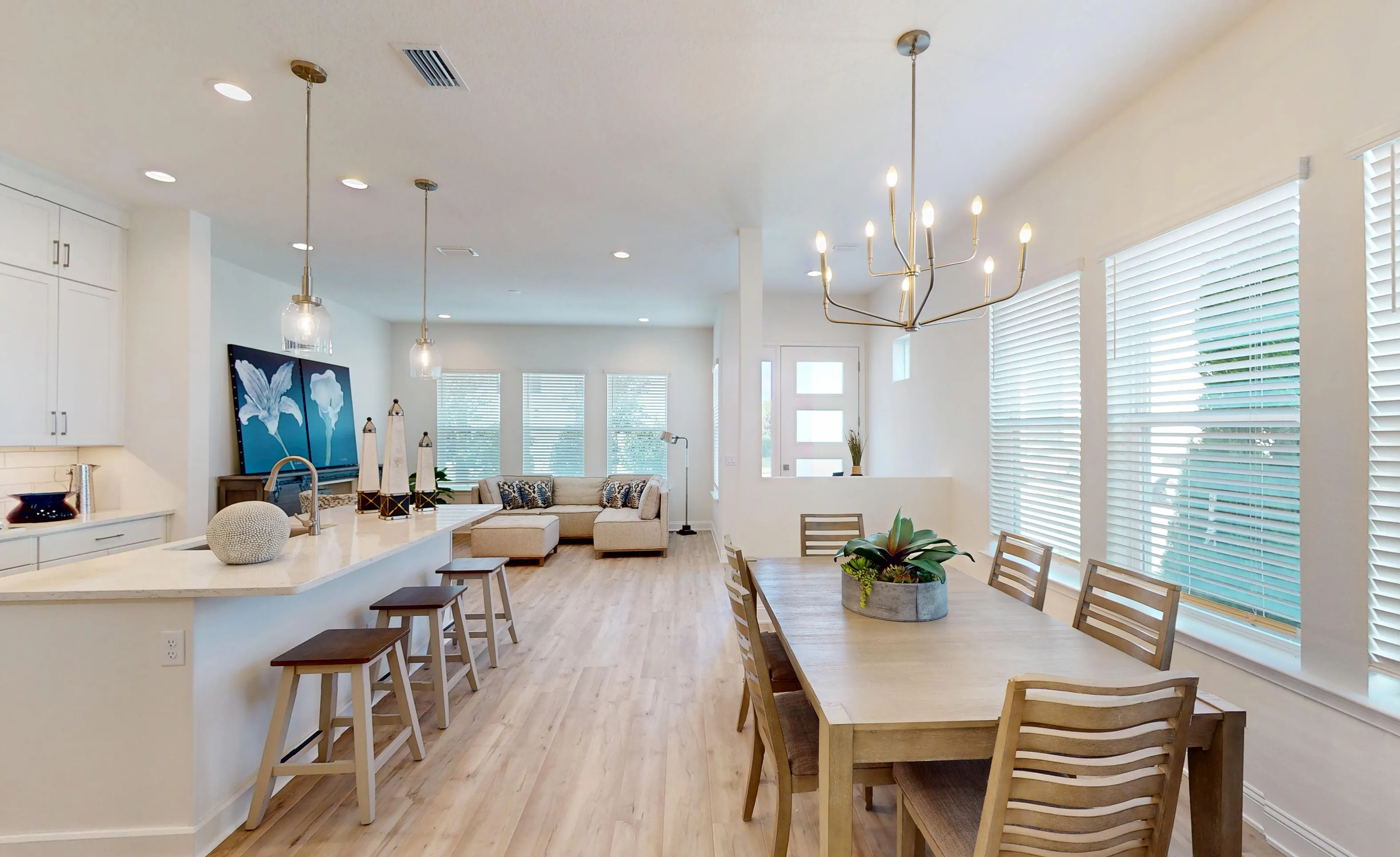 Dining room and open kitchen with a wooden table, island with stools, light-colored flooring, and large windows with blinds.