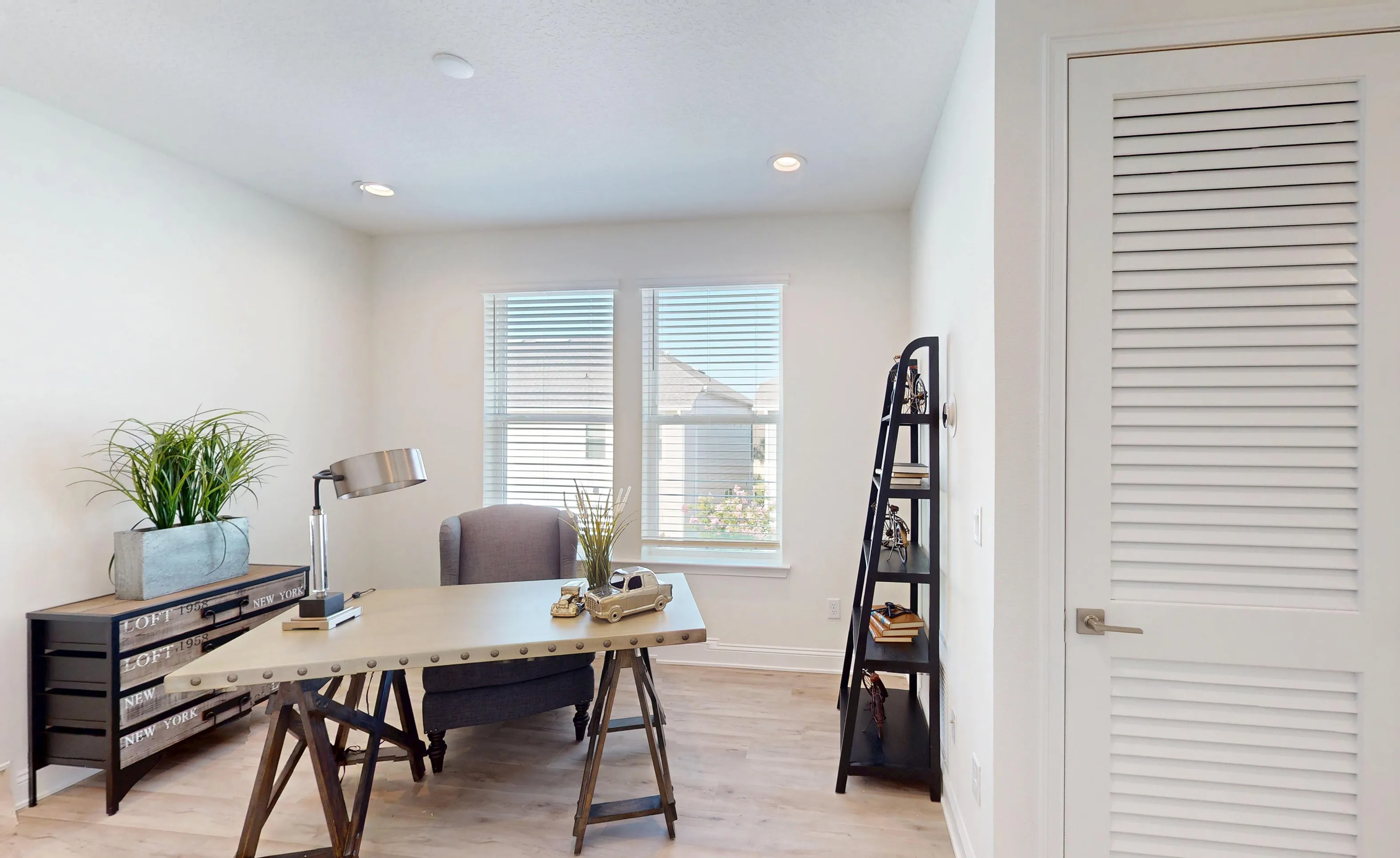 Home office in the Bartram B plan featuring a metal-topped desk, grey armchair, white walls, and louvered closet door.