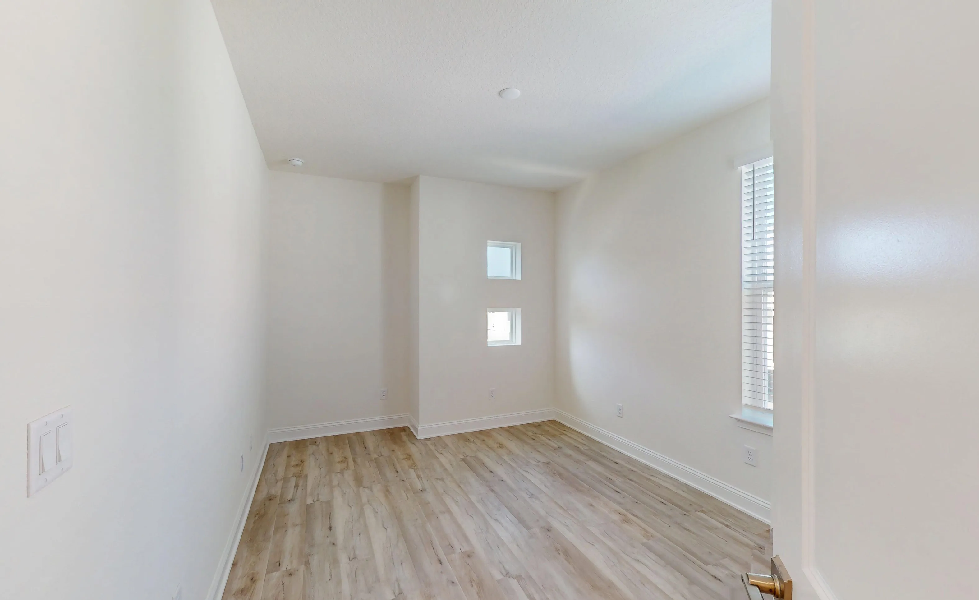 Secondary bedroom with light wood-look flooring, white walls, a large window with blinds, and two small square windows.
