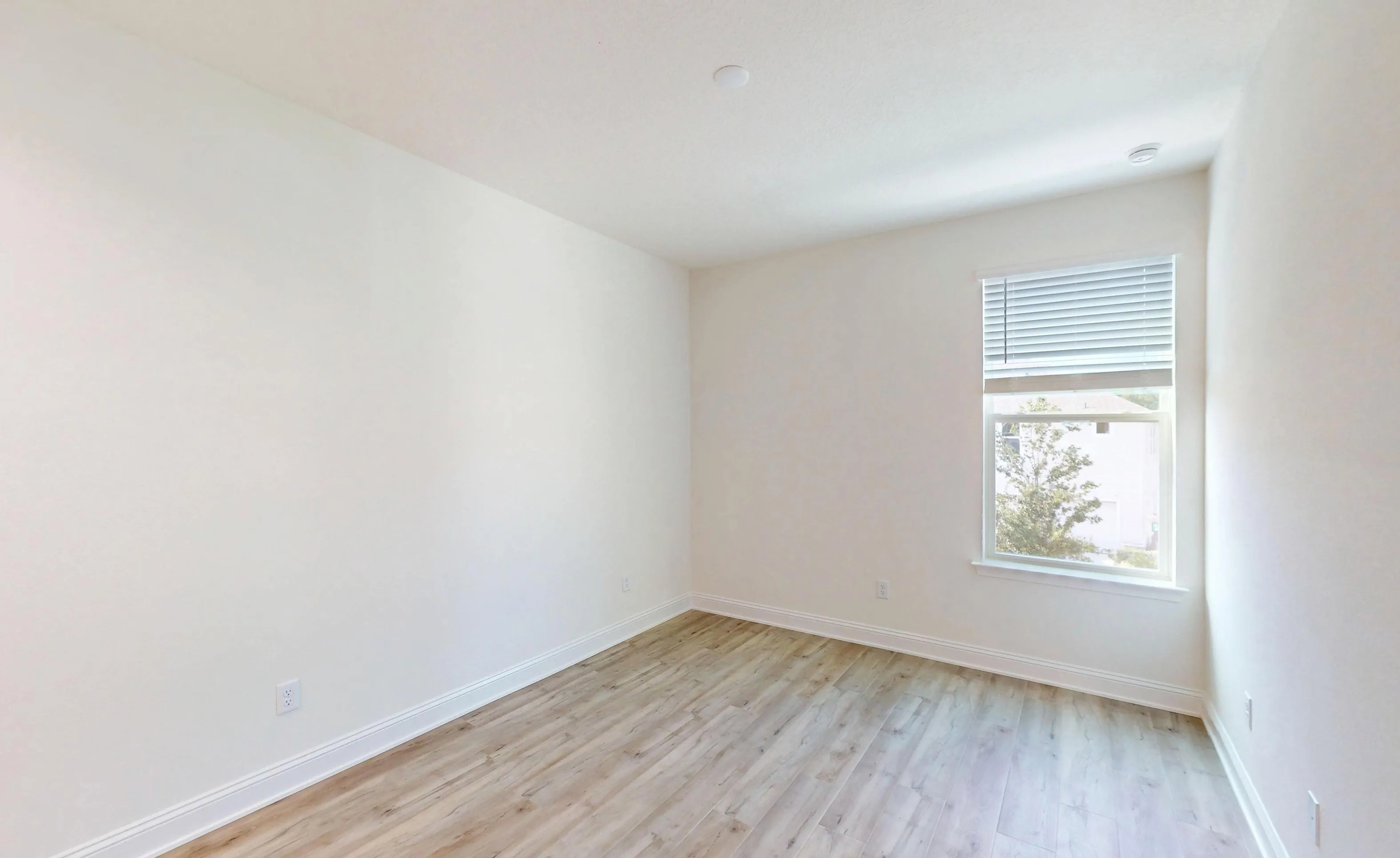 Empty secondary bedroom with light wood-look flooring, white walls, white baseboards, and a window with blinds.