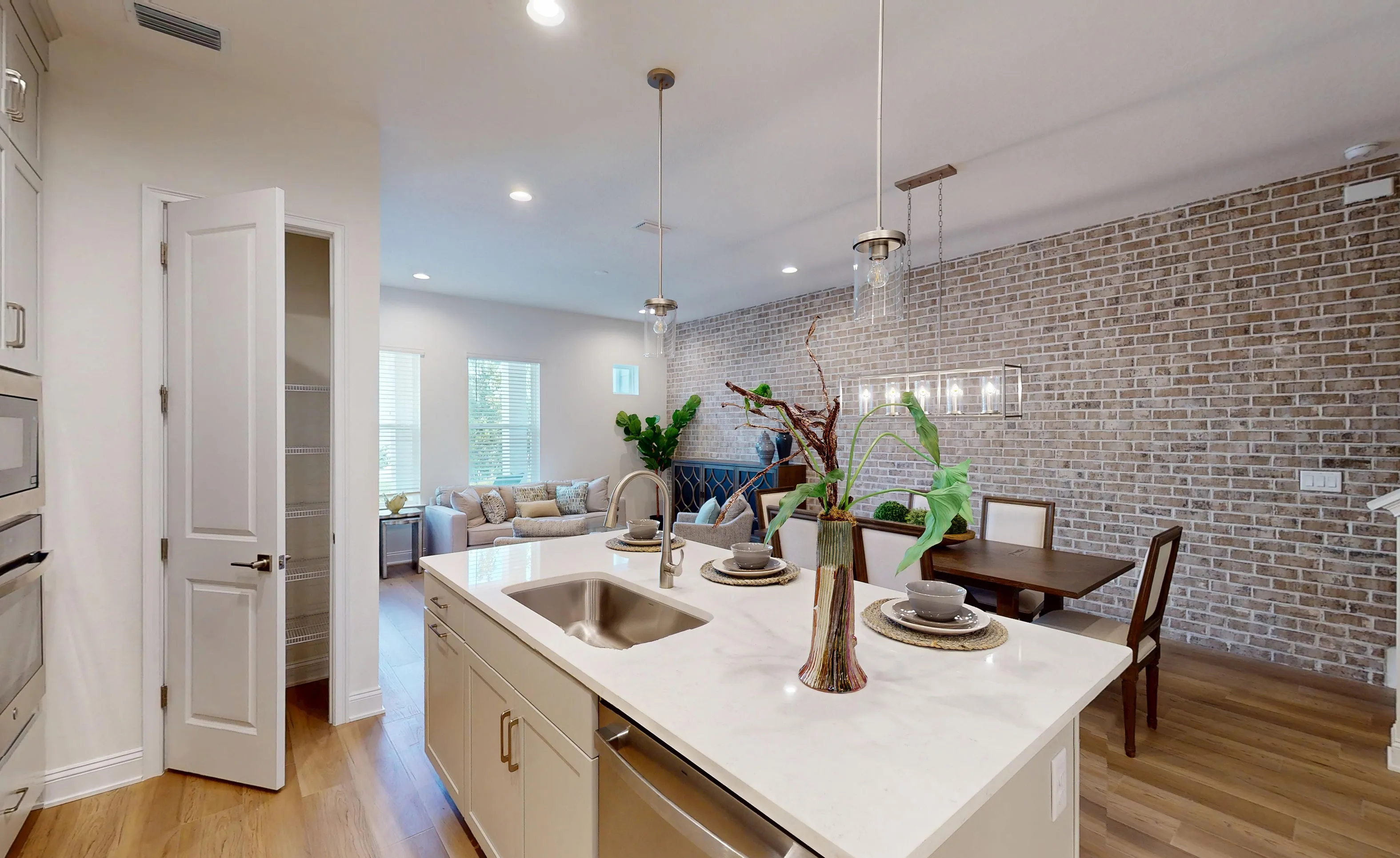 Kitchen island with a sink and white countertop, with an open pantry door and a dining area featuring a brick accent wall.