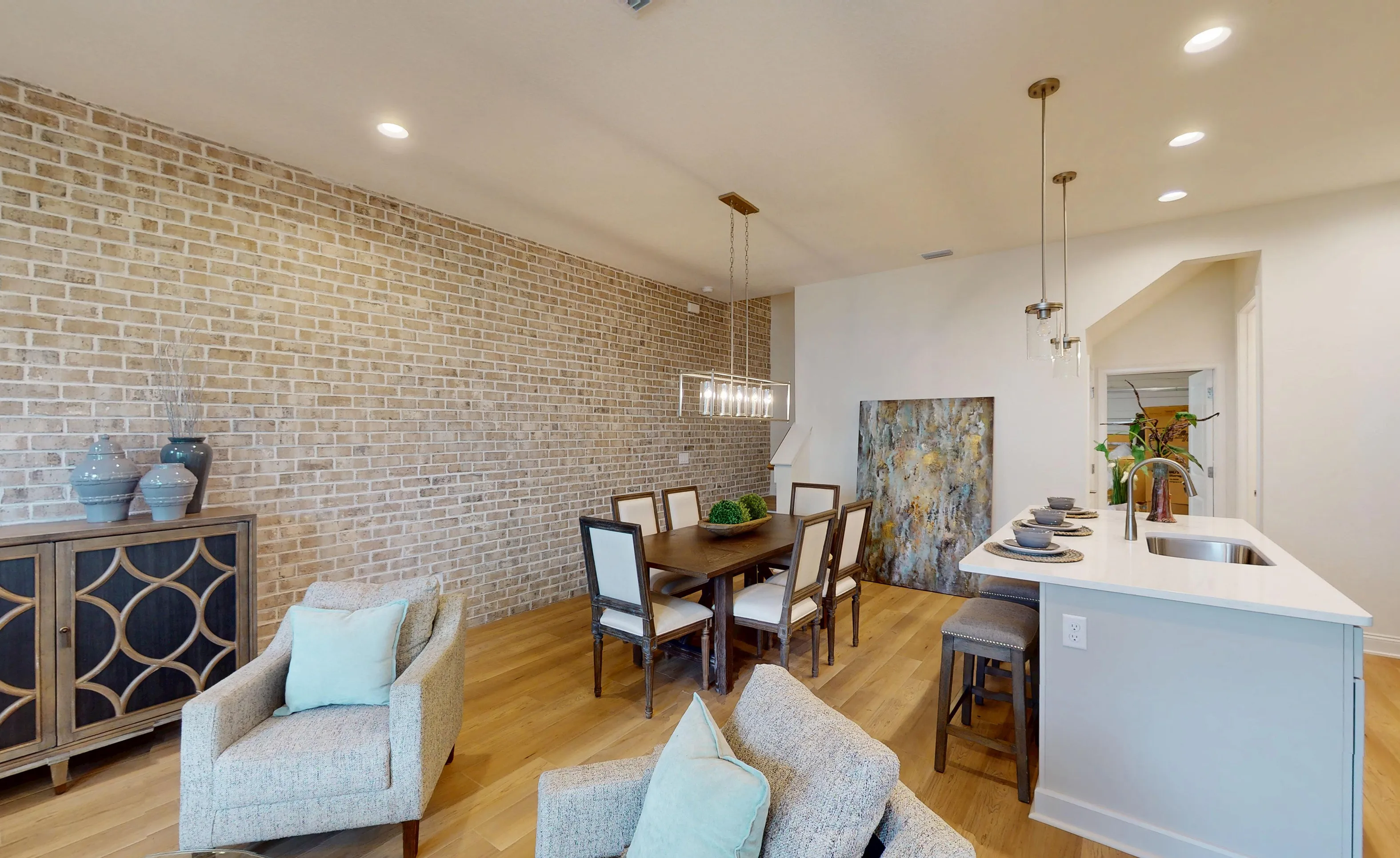 Dining room with hardwood floors, light brick accent wall, wooden table with six chairs, and adjacent kitchen island.