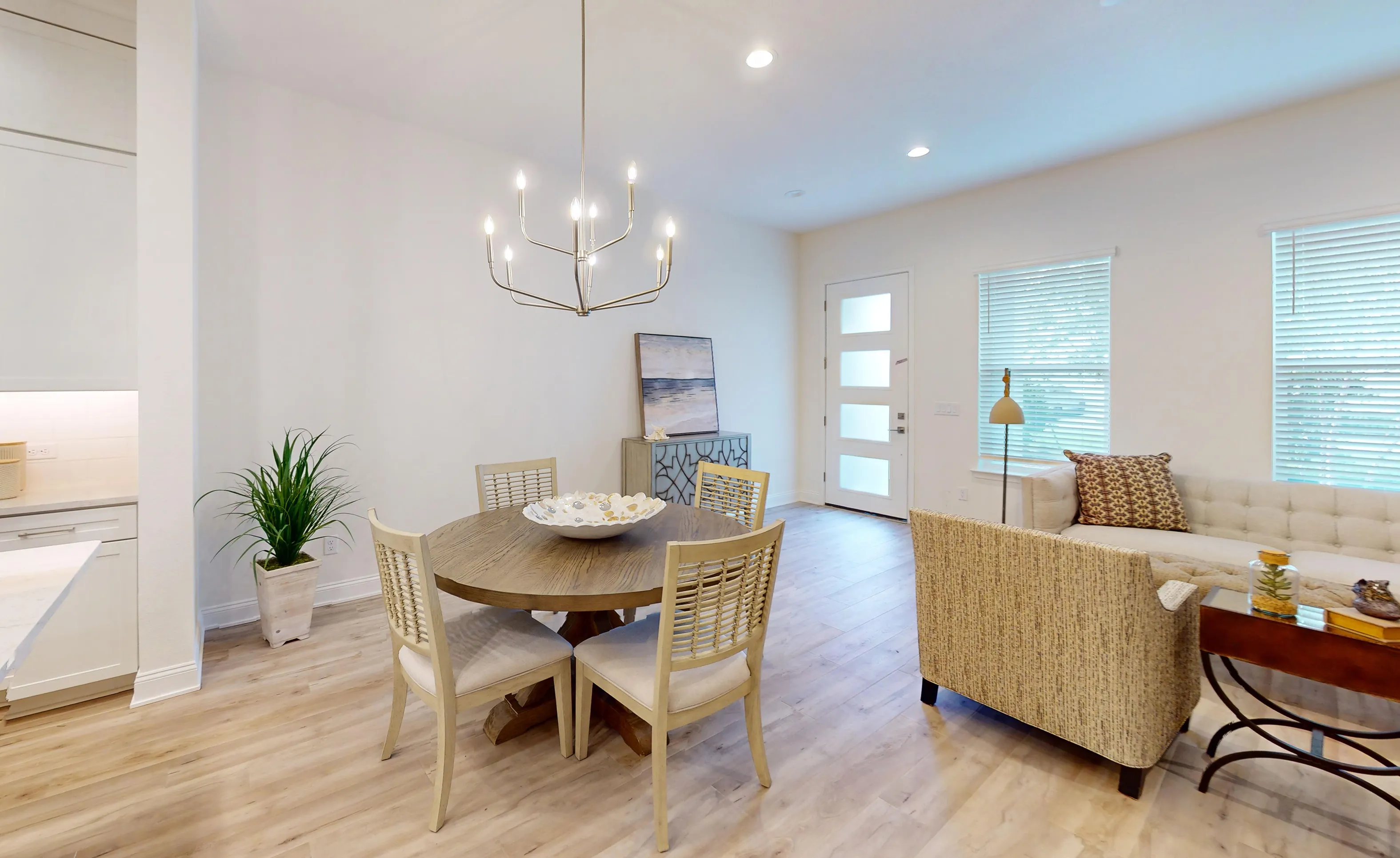 Dining room of the Deleon-D plan with a round wooden table, chandelier, and adjacent living area with wood-look flooring.