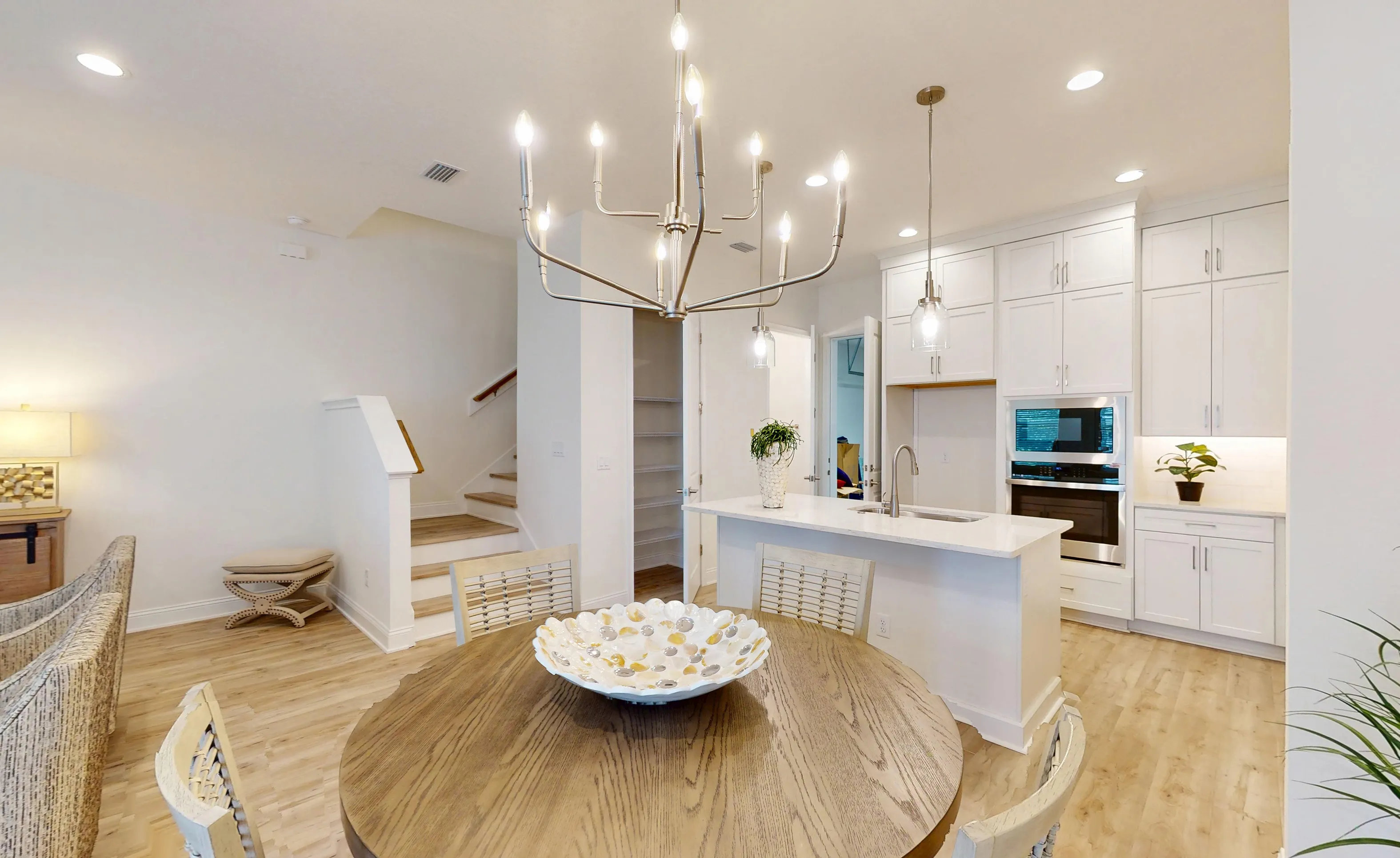 Breakfast nook with a round wooden table, chandelier, and adjacent kitchen with white cabinets, island, and wall oven.