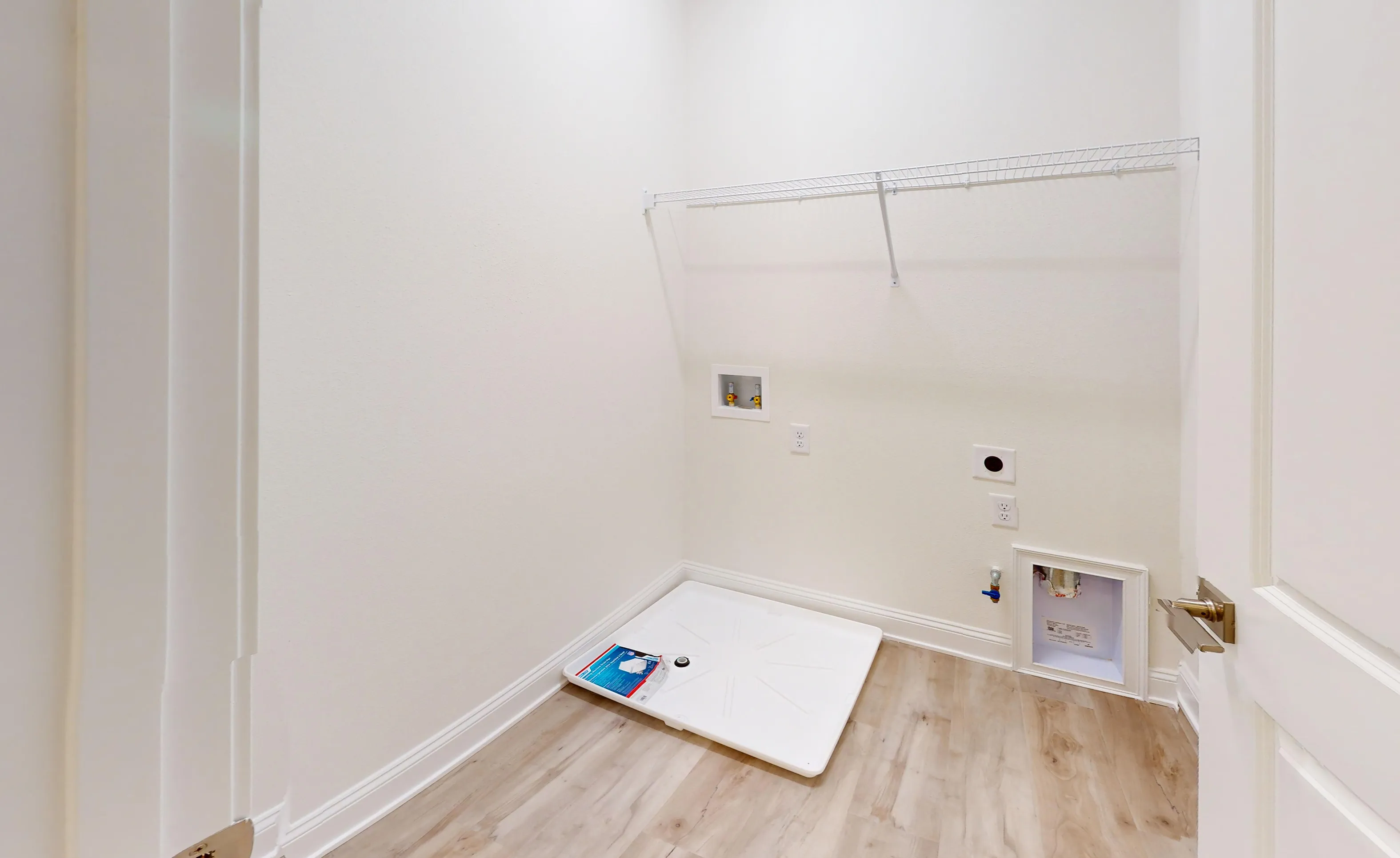 Laundry room with a white floor drain pan, utility hookups, and a wire shelf on a white wall above wood-look flooring.