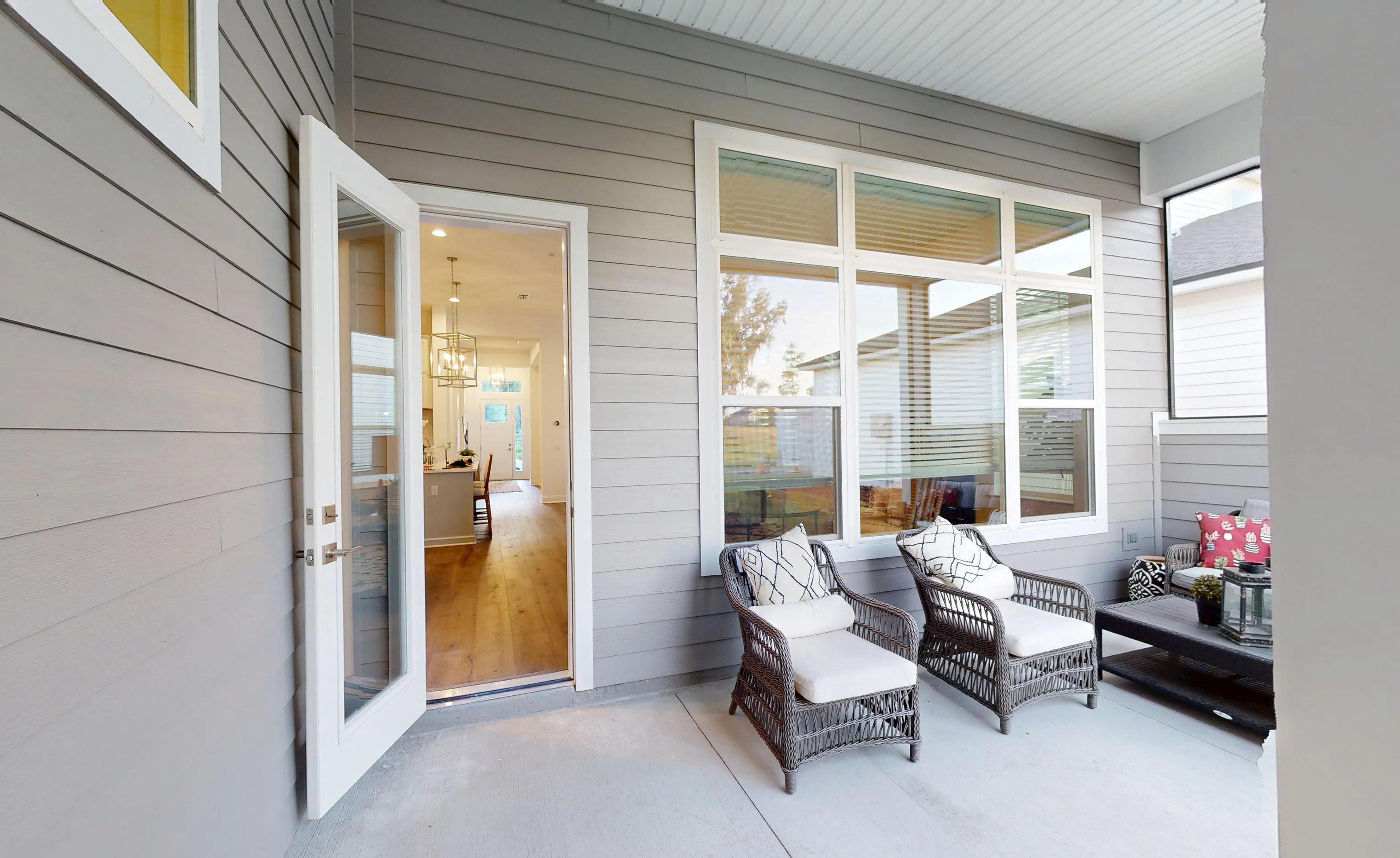Covered porch with grey horizontal siding, concrete floor, two wicker chairs, and a glass door opening to the interior.