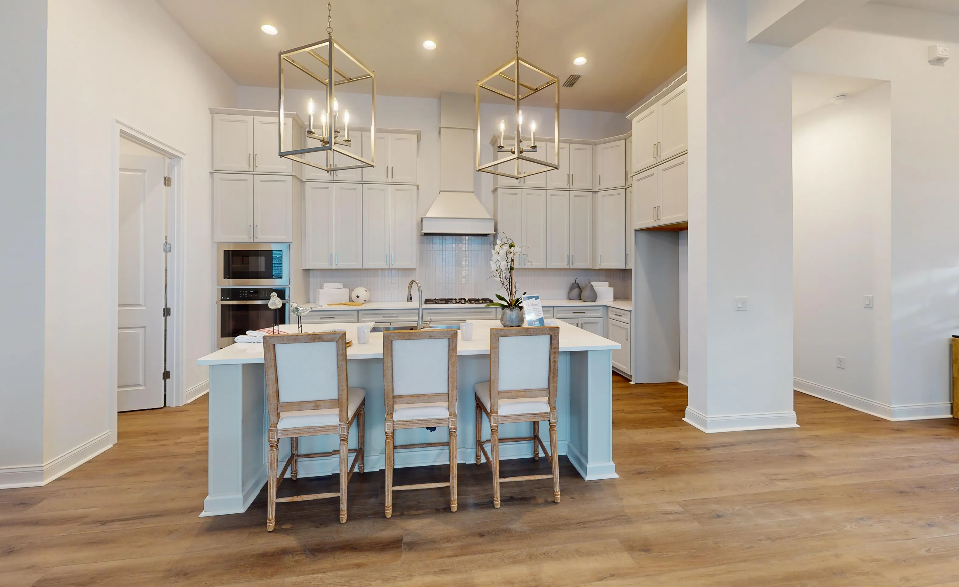 Kona kitchen in Oakmont with white cabinets, large island with three chairs, wood flooring, and two geometric lanterns.