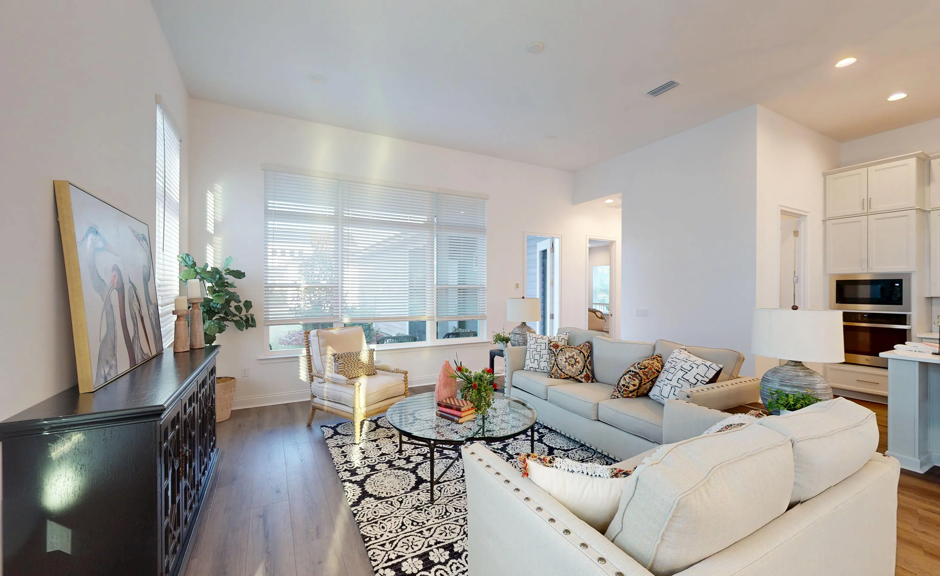 Kona plan living room with light furniture, a round glass coffee table, patterned area rug, and wood-look flooring.