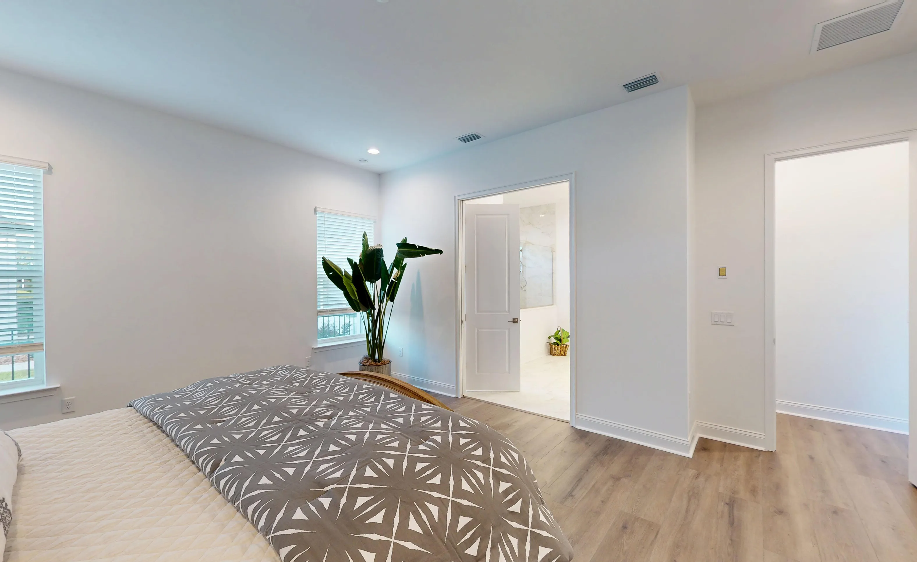Primary bedroom of the Kona plan with wood-look flooring, white walls, two windows, and an en-suite bathroom entrance.