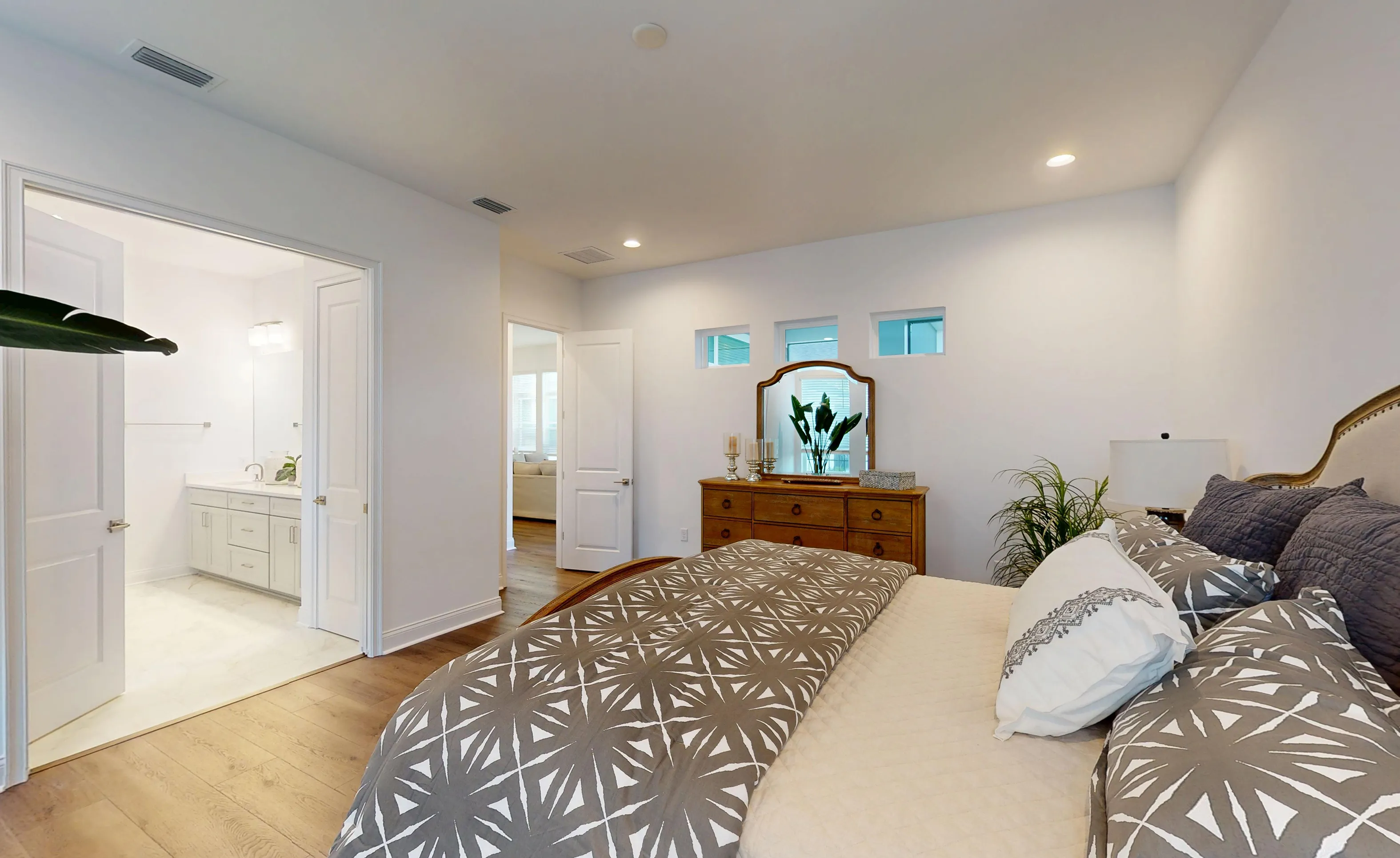 Primary bedroom of the Kona plan featuring wood-look flooring, a wood dresser with mirror, and an en suite bathroom.
