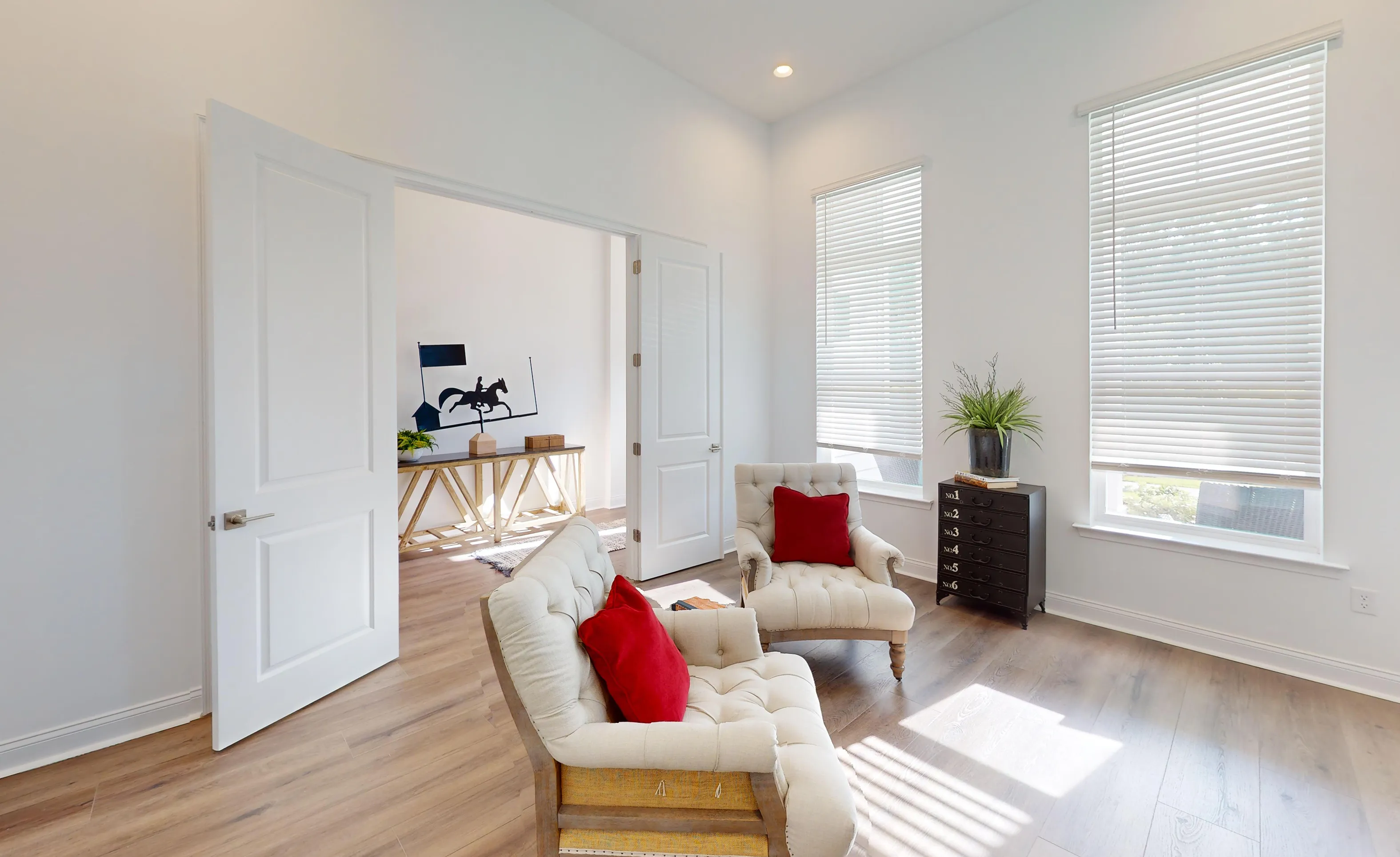 Home office in the Kona plan with white walls, wood-look flooring, two cream tufted chairs with red pillows, and dual doors.