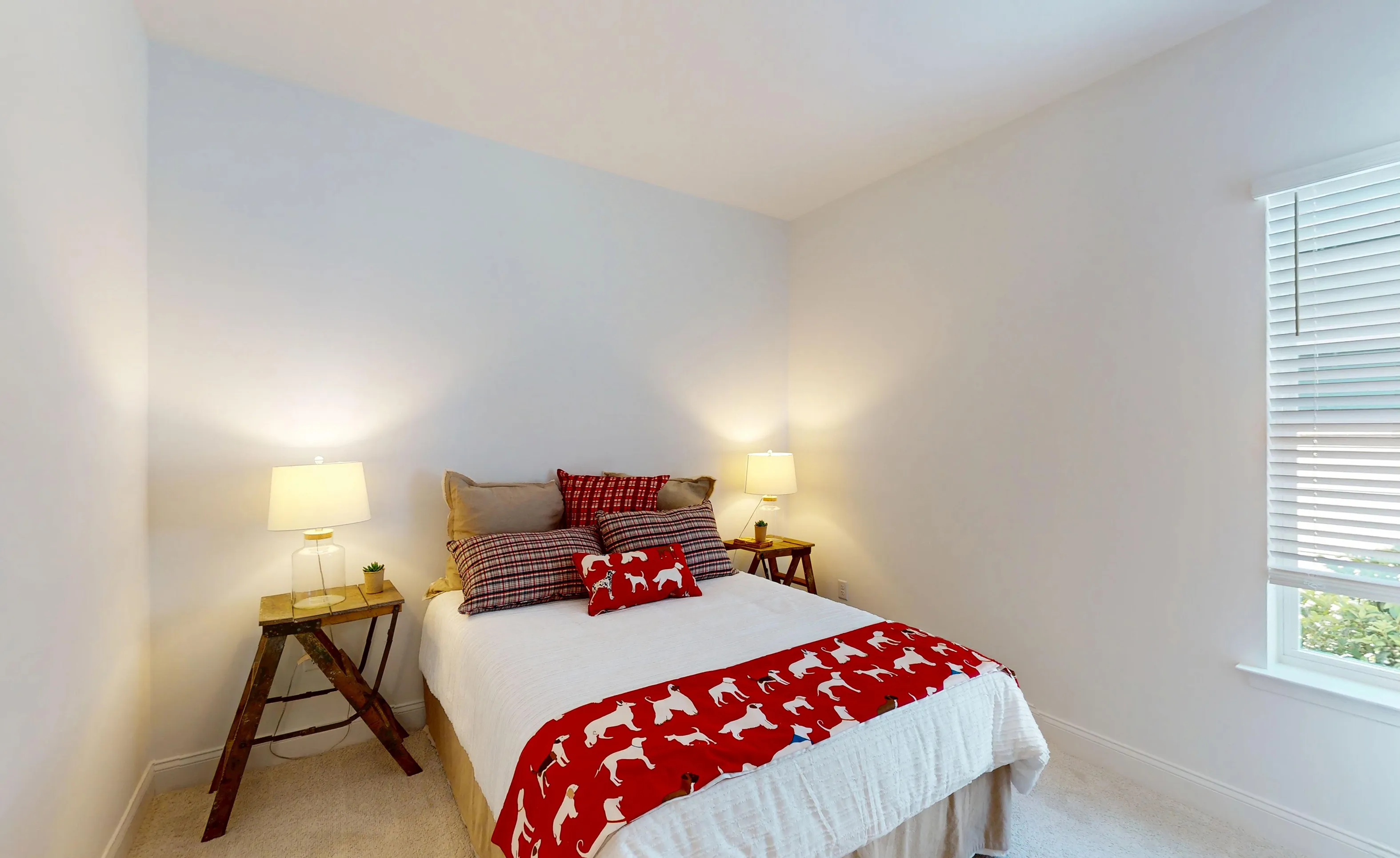 Secondary bedroom in the Kona plan featuring a queen bed with a red dog-print runner, bedside lamps, and a window with blinds.