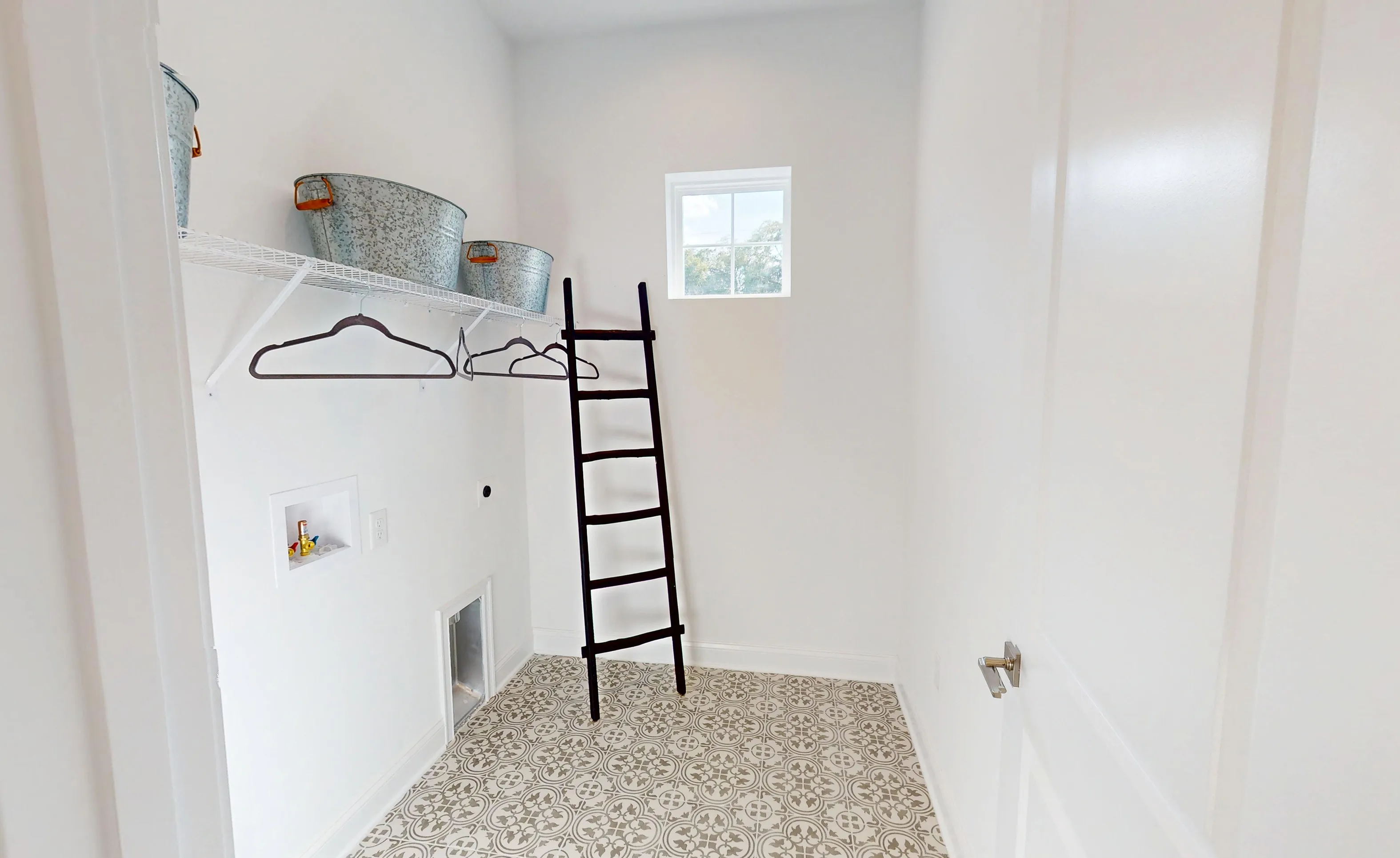 Laundry room in the Kona plan with white walls, patterned floor tiles, wire shelving, and a black decorative ladder.