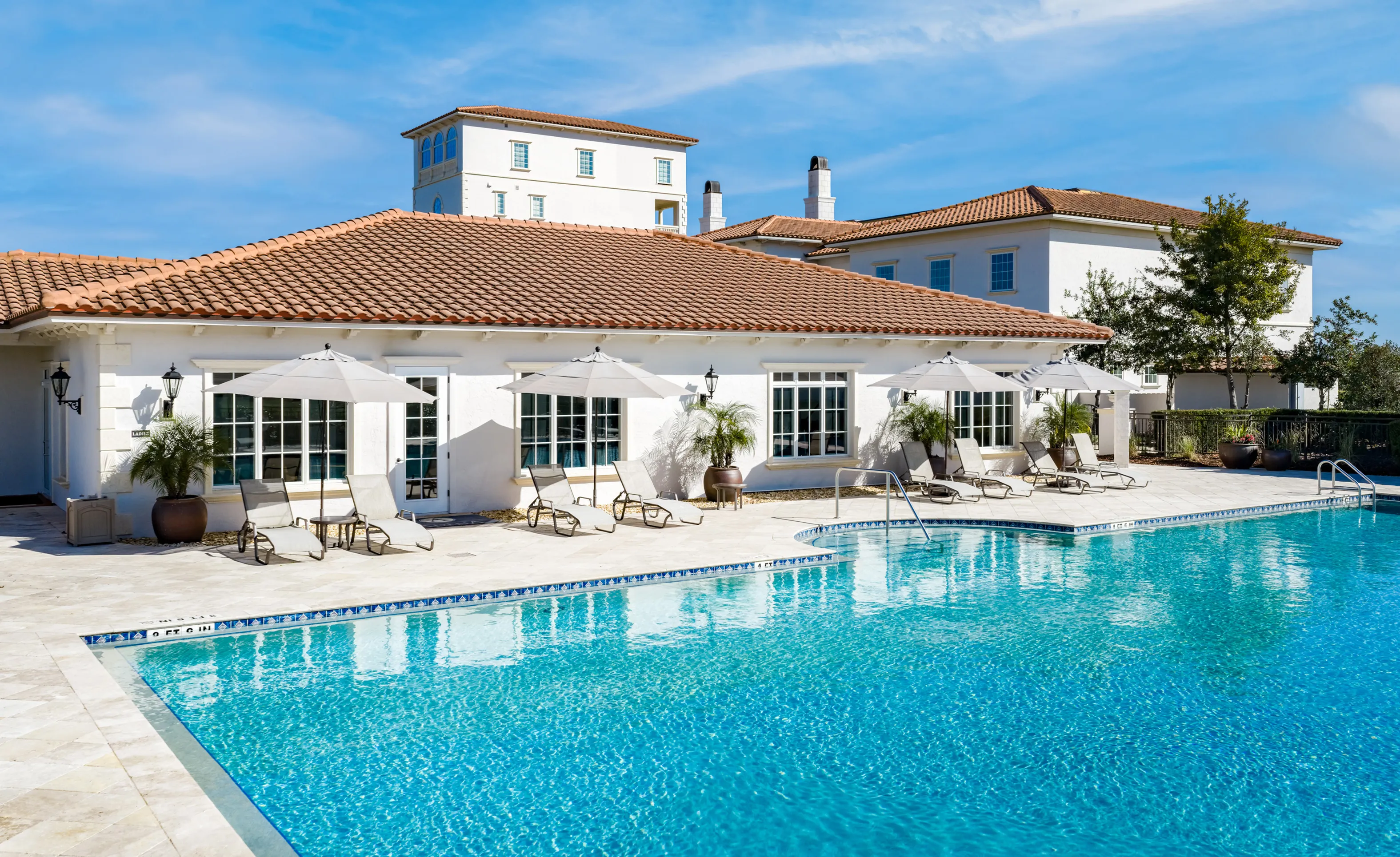 Swimming pool area at IronCrest community featuring a paved deck, lounge chairs, umbrellas, and white buildings.