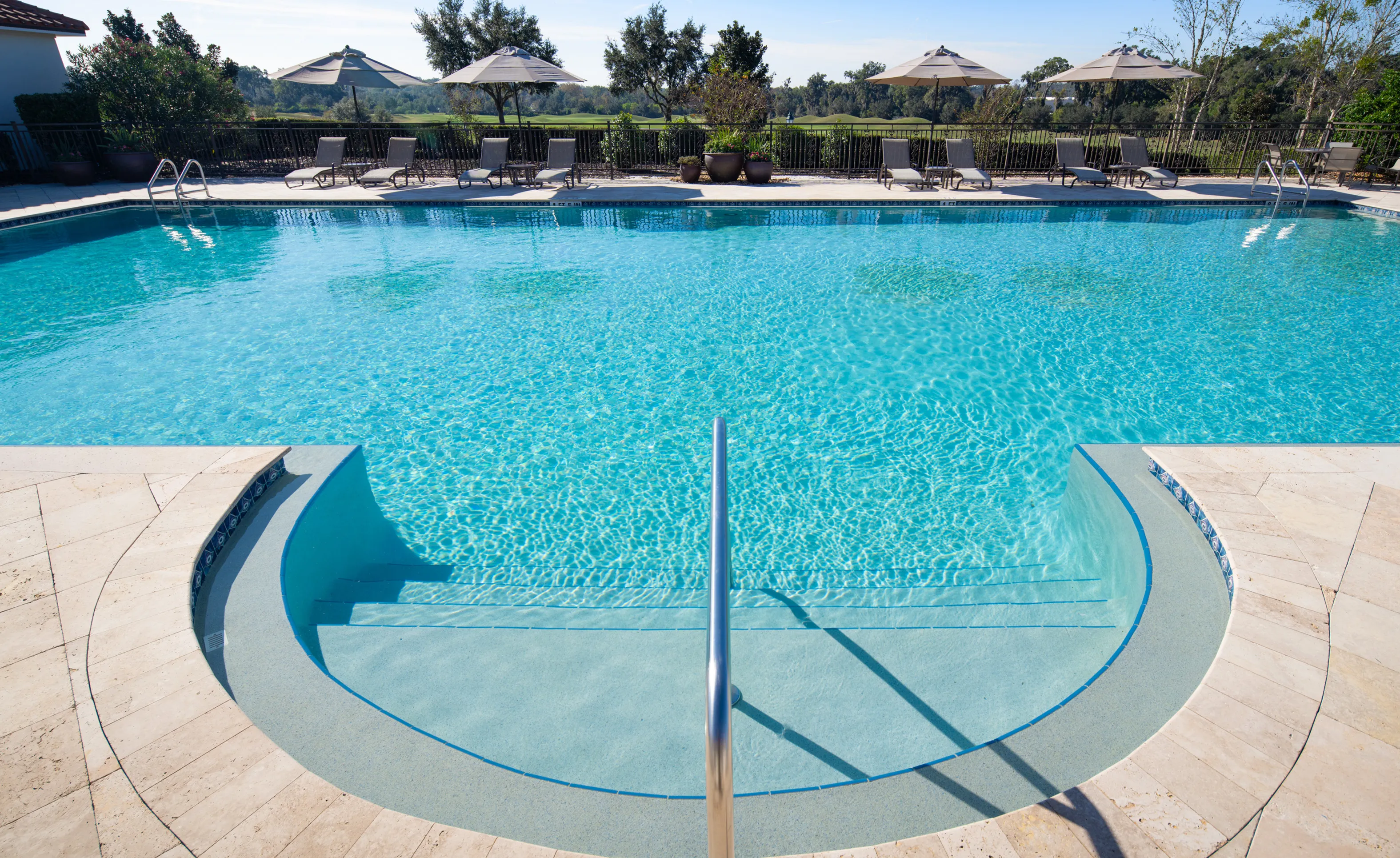 Swimming pool with curved stairs and a metal handrail at IronCrest, featuring lounge chairs under umbrellas on the deck.