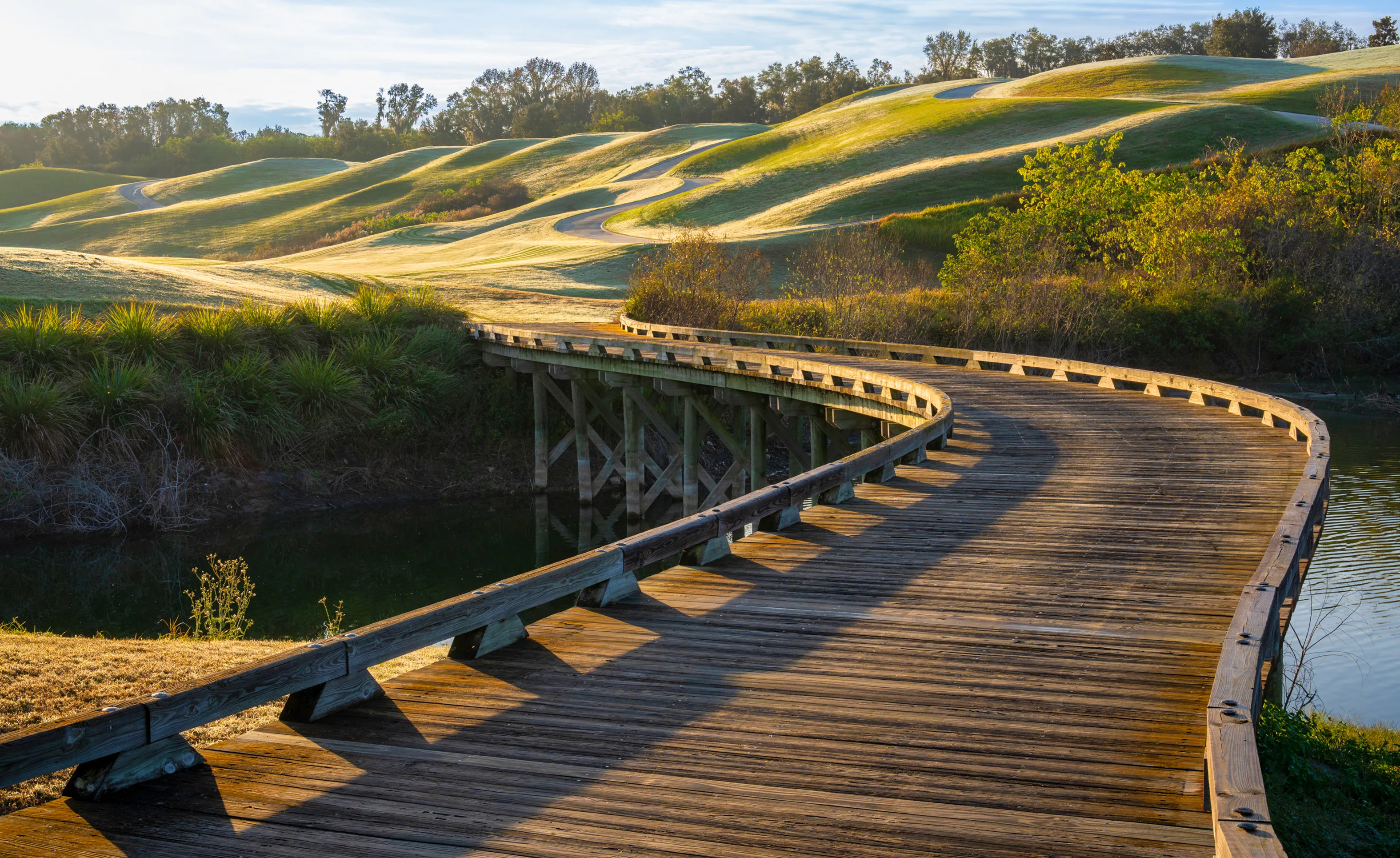 Wooden footbridge over a pond leading to rolling green hills at IronCrest community.