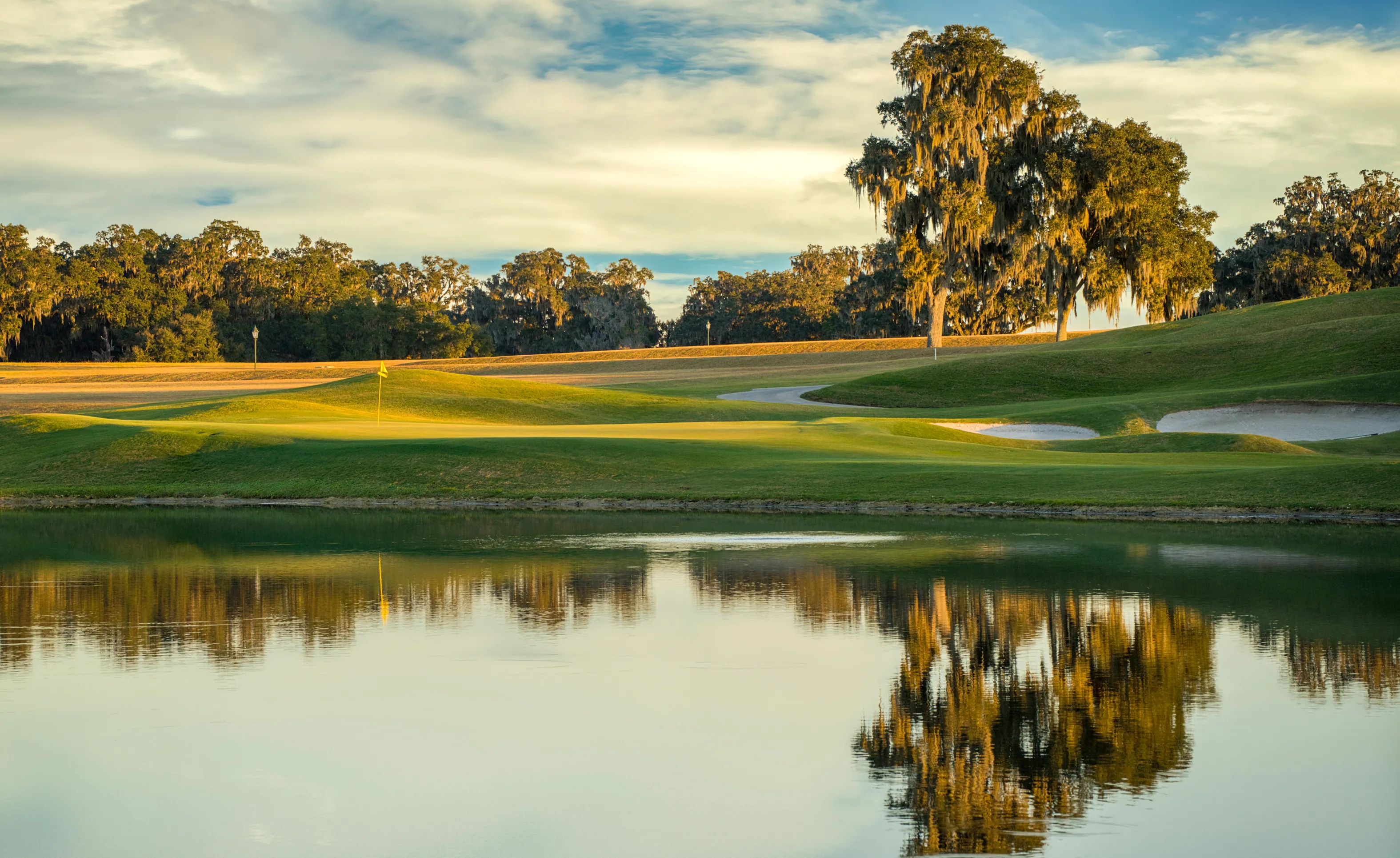 Golf course view at IronCrest featuring a green, sand bunkers, and a lake reflecting large moss-draped oak trees at sunset.
