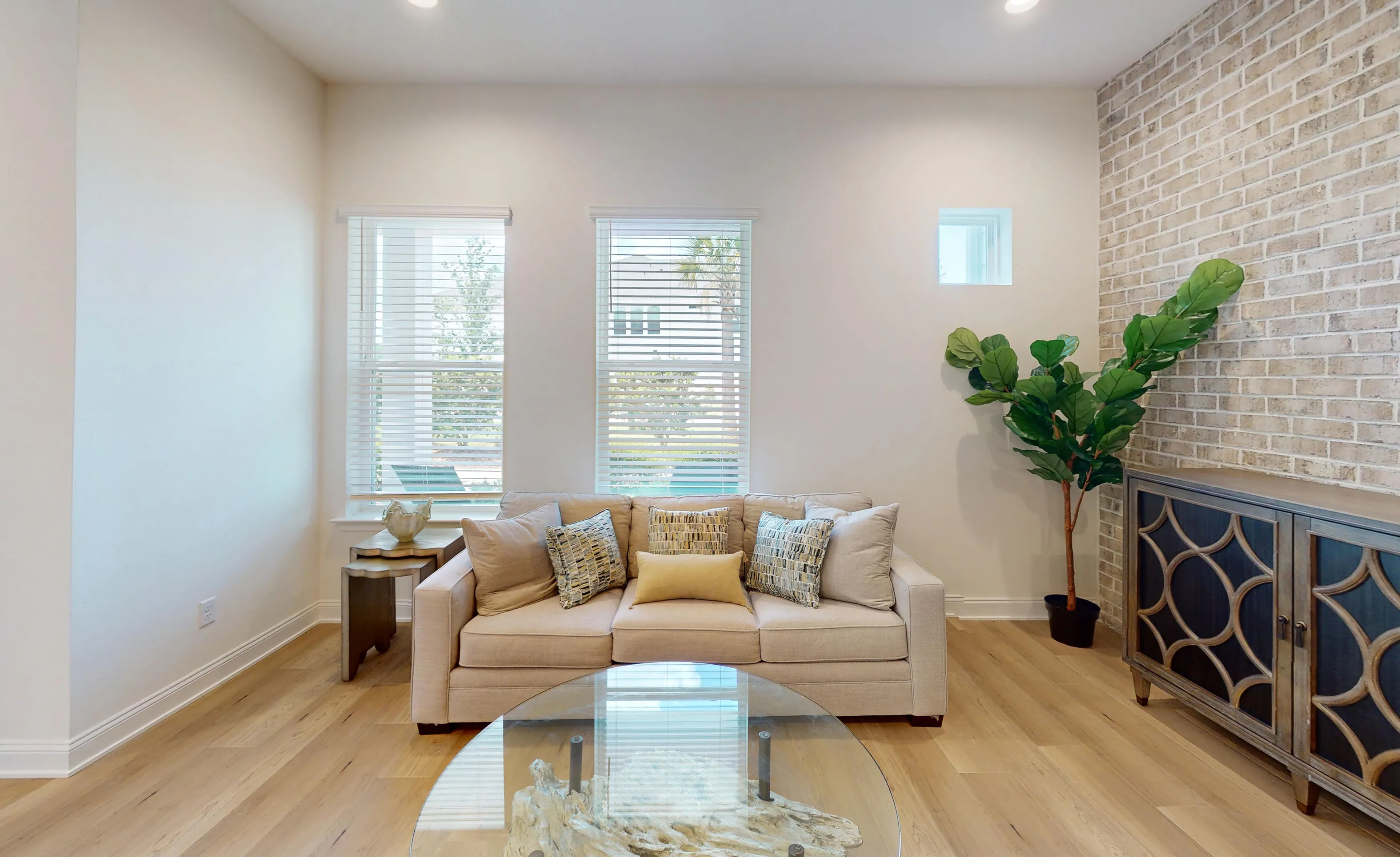 Living room in the Castillo plan with light wood floors, a beige sofa, two windows, and a white brick accent wall.