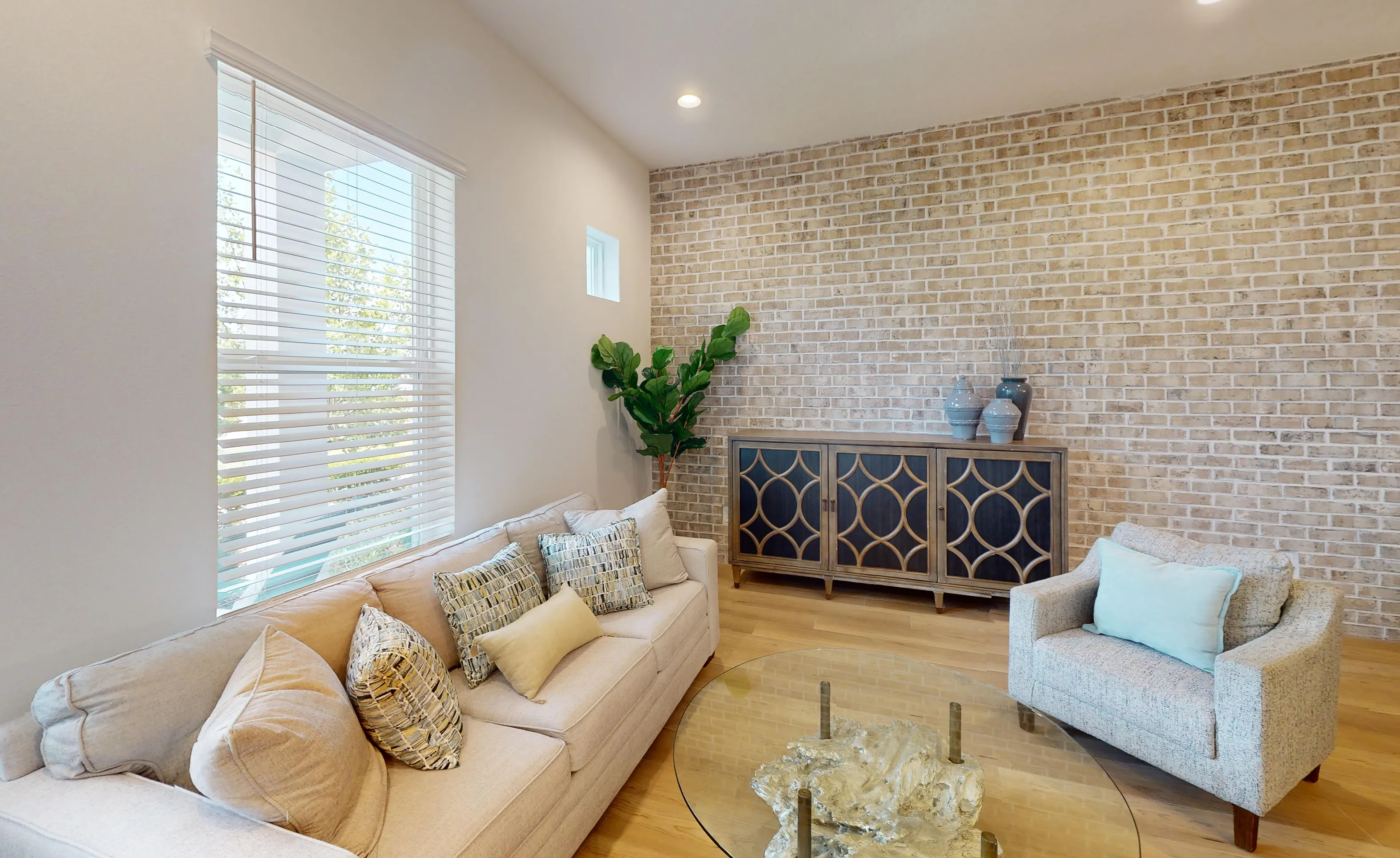 Living room in Castillo plan with light beige brick accent wall, wooden floors, beige sofa, and glass coffee table.