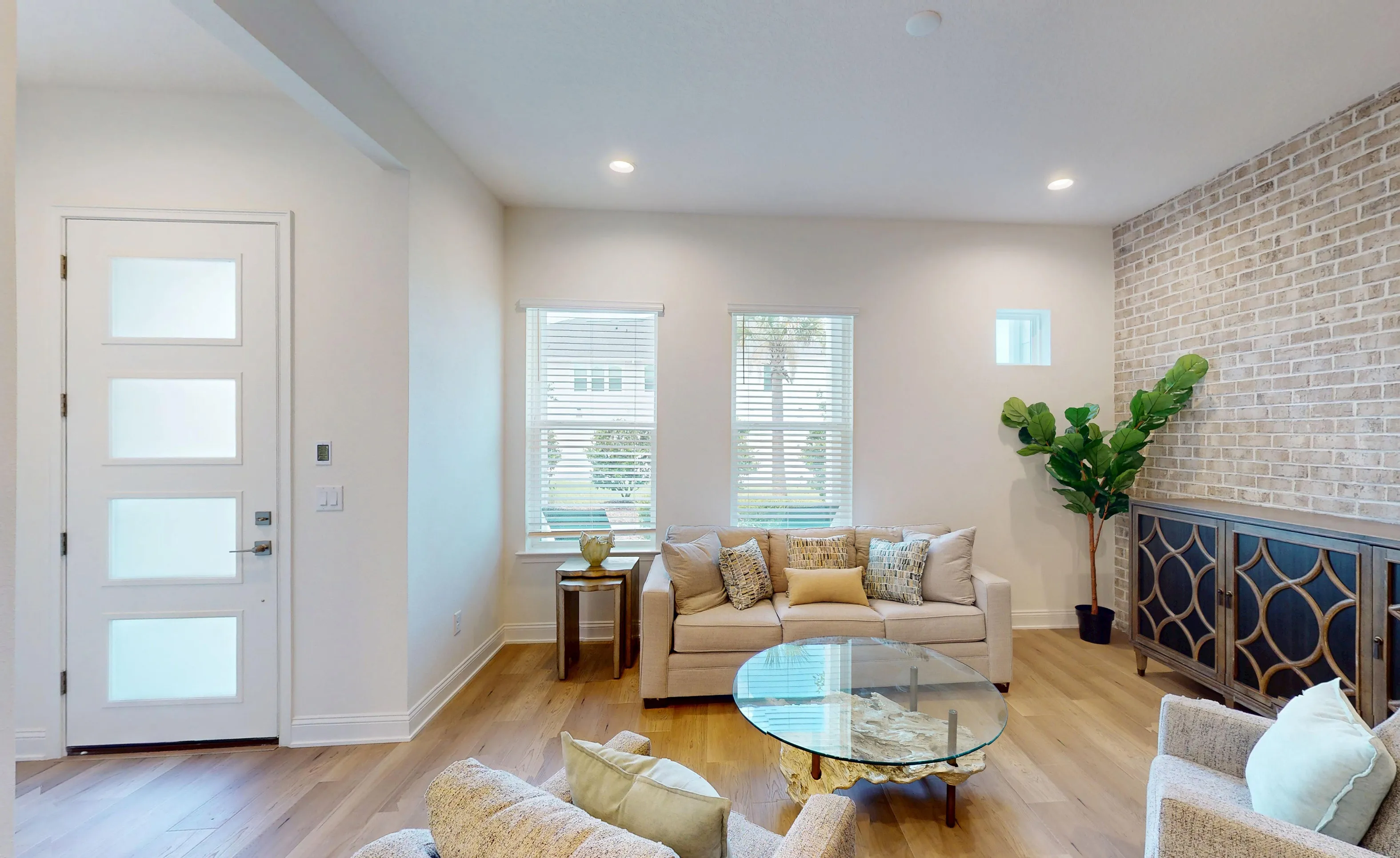 Living room of the Castillo C plan with light wood floors, a brick accent wall, and a white multi-panel entry door.