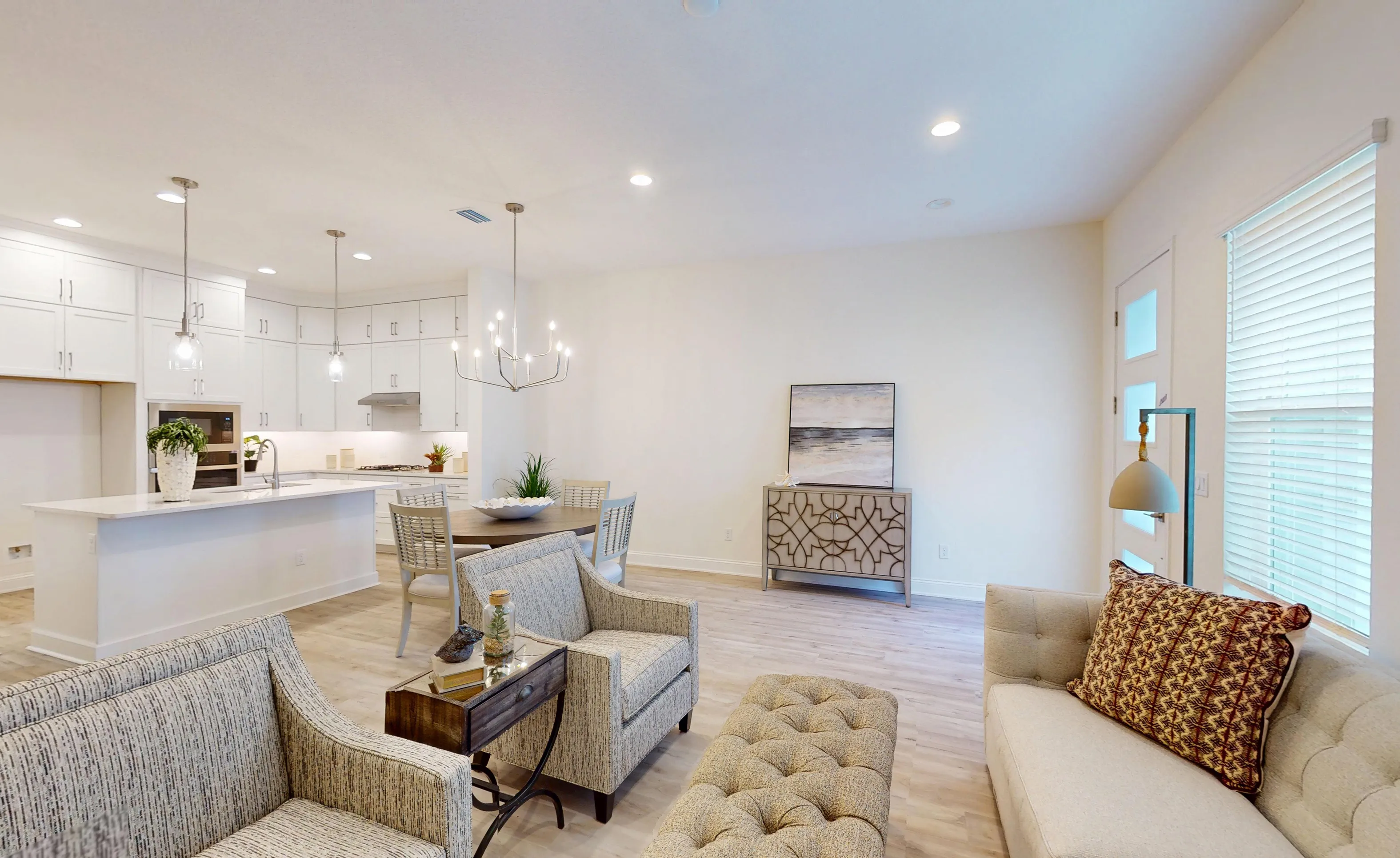 Open-concept living room and kitchen in the Deleon D model, featuring wood-look flooring, light furniture, and white cabinets.