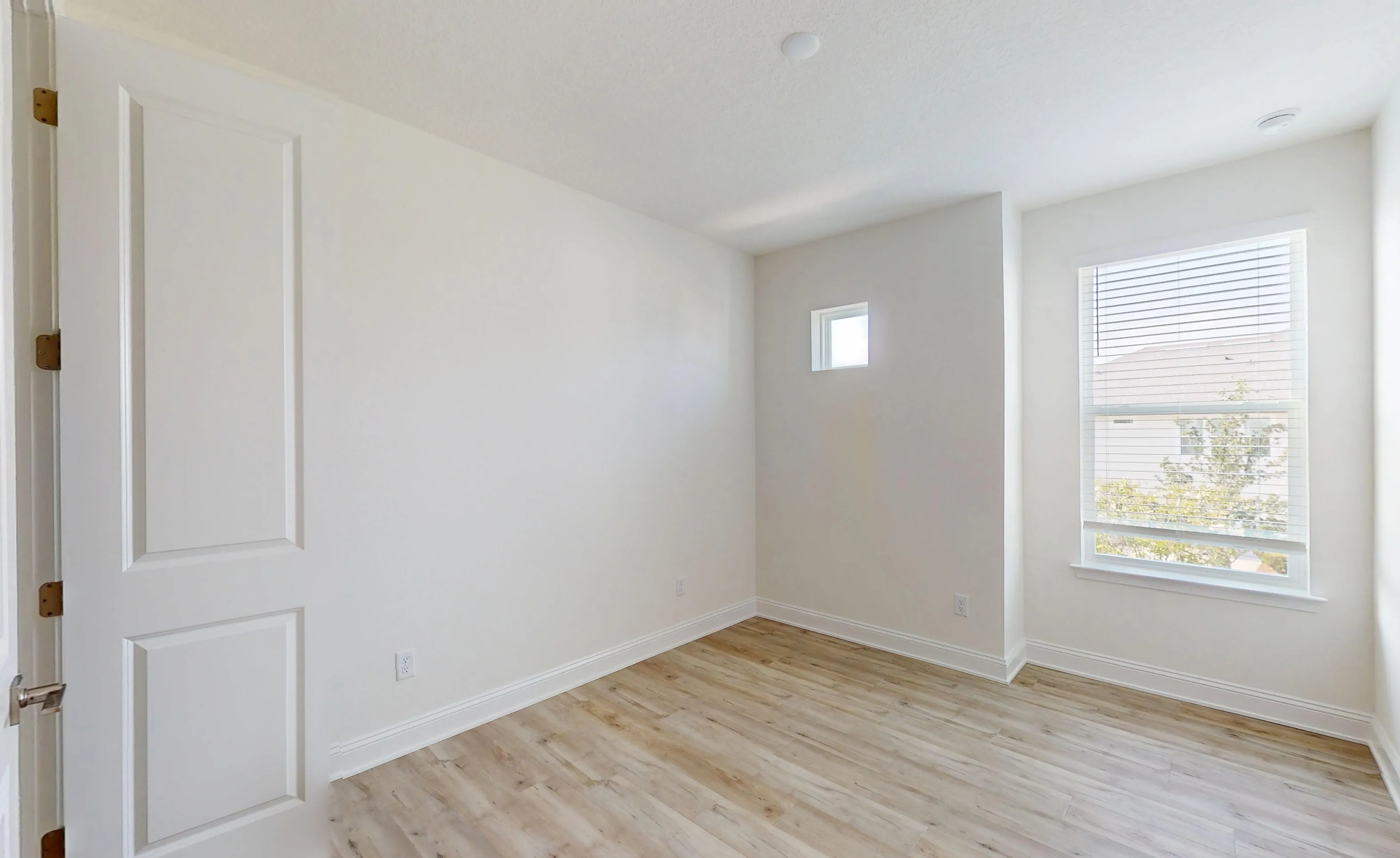 Secondary bedroom with light wood-style flooring, white walls, a large window with blinds, and a small square transom window.
