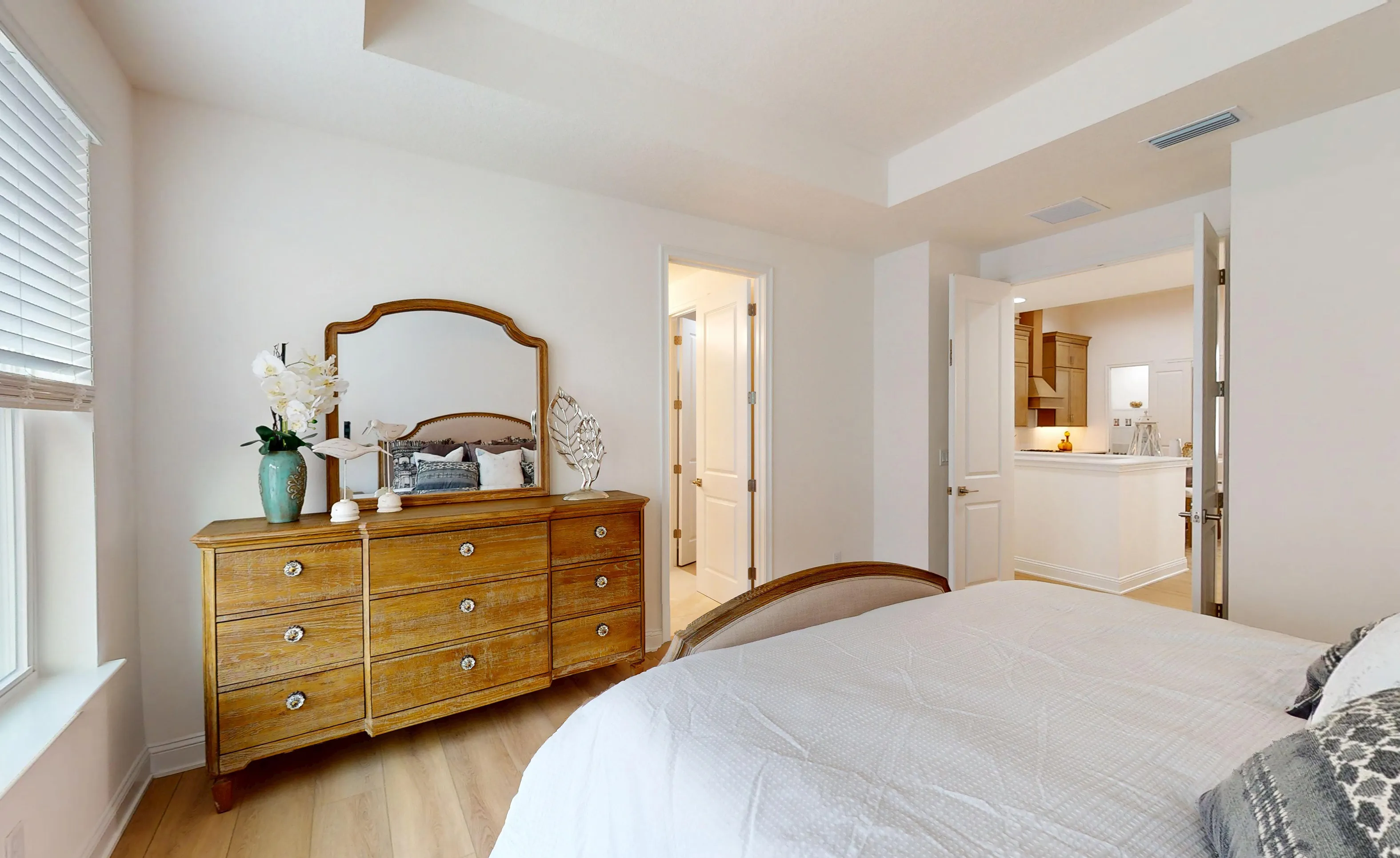 Primary bedroom of the Gulf plan with light wood flooring, a wooden dresser with a mirror, and an open doorway to a kitchen.