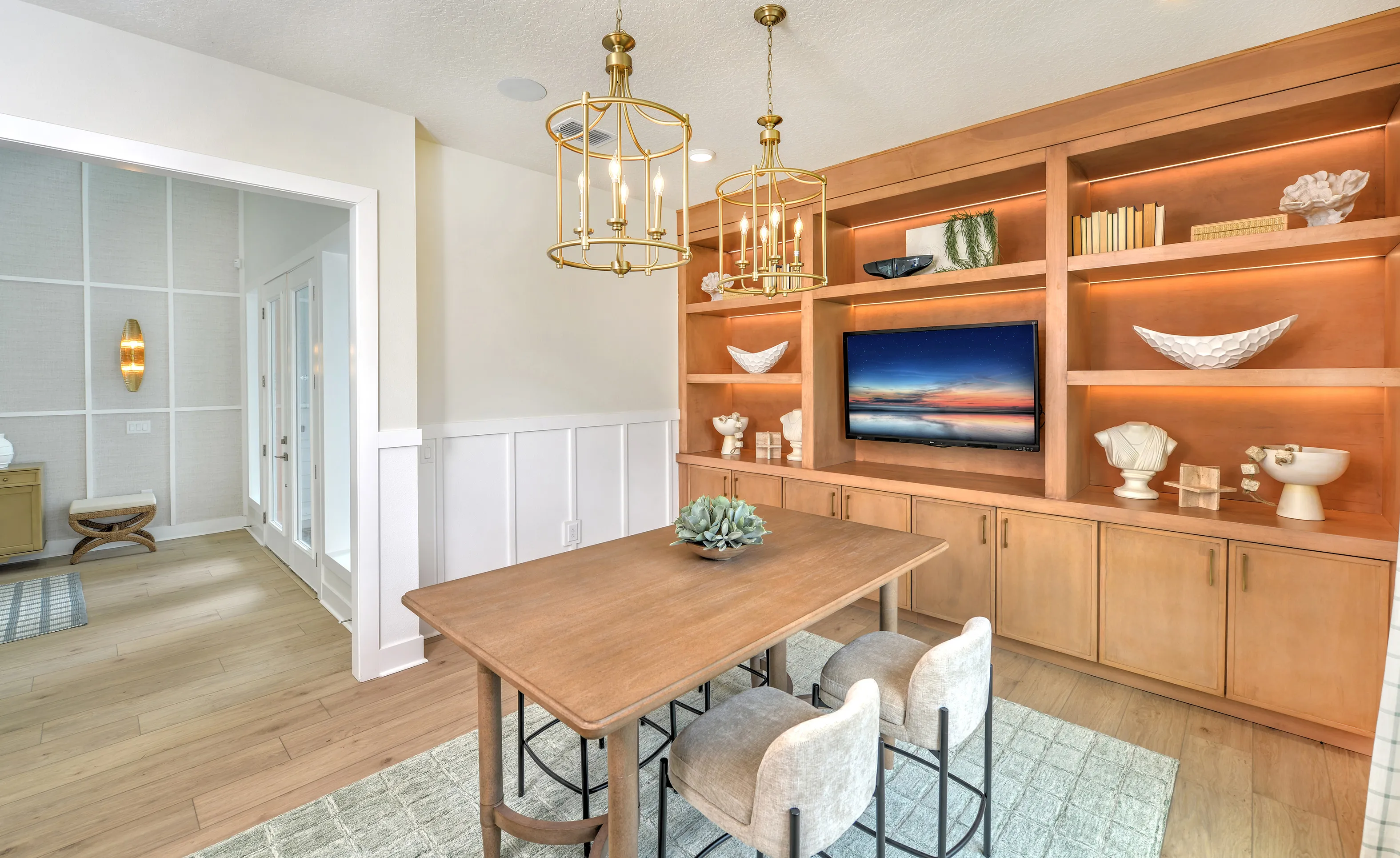 Study with wood table, two chairs, brass pendant lights, and built-in wood shelving featuring a mounted television.