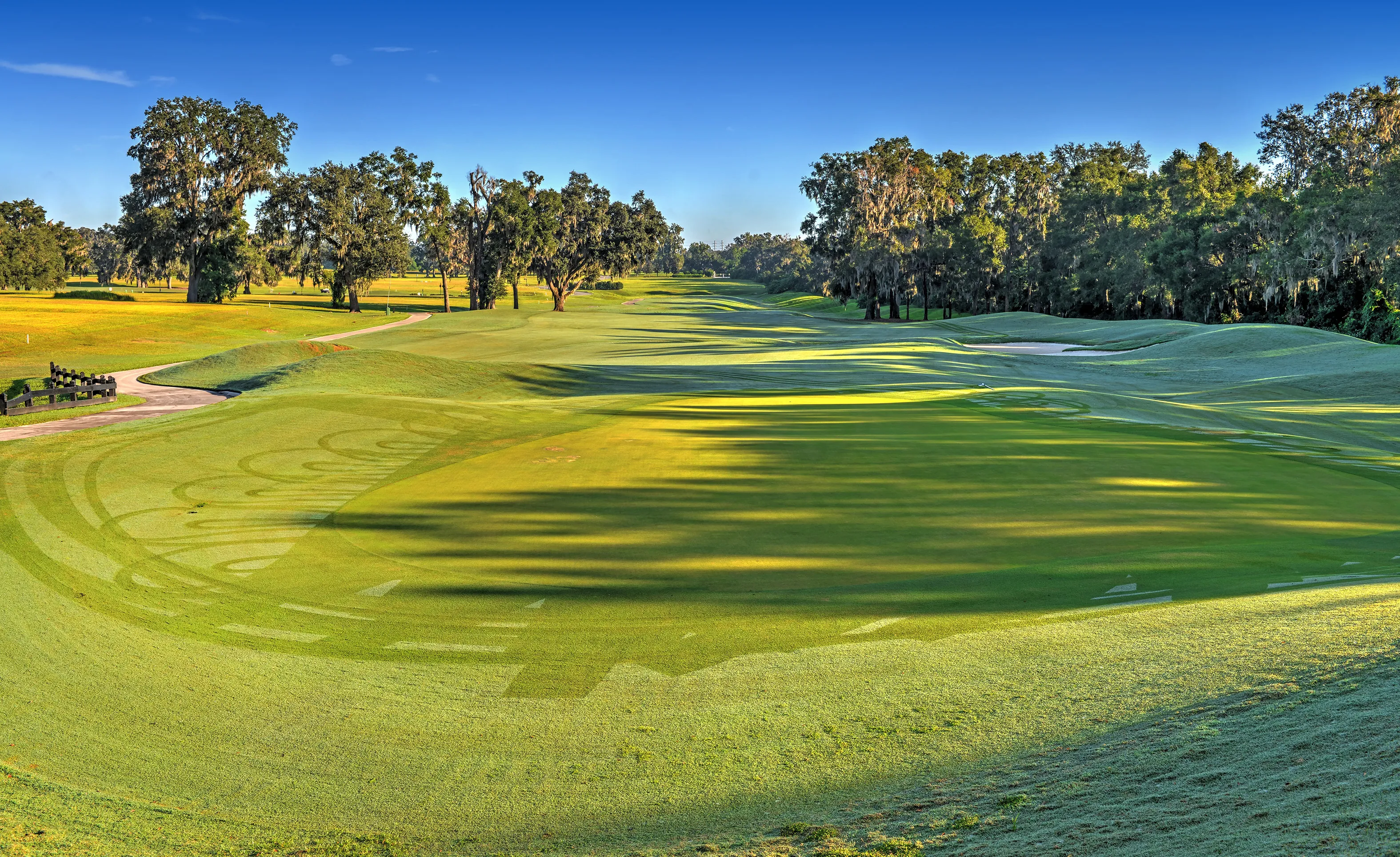 A panoramic view of an IronCrest golf course fairway with lush green grass, rolling hills, and mature trees under a blue sky.
