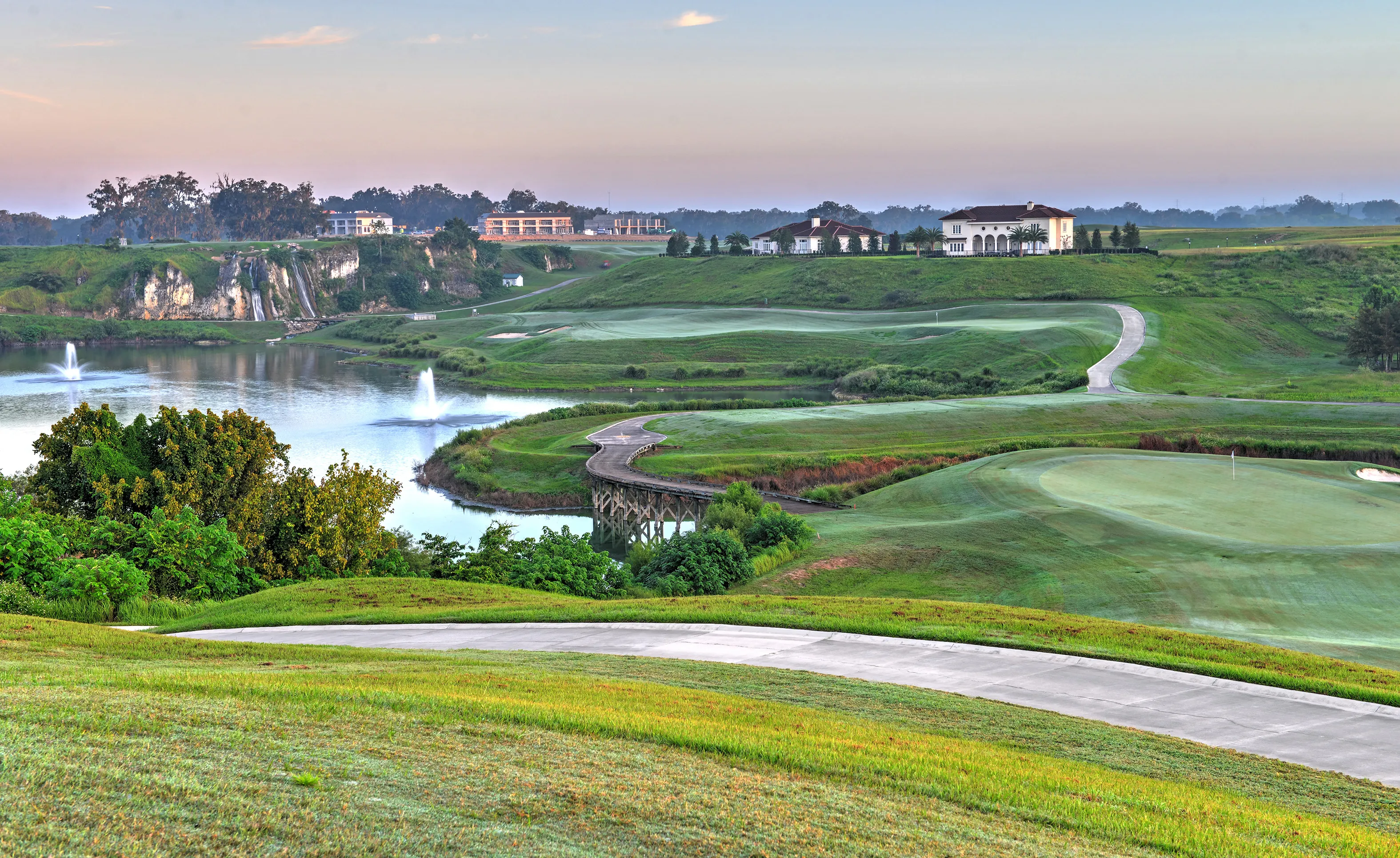 Golf course view at IronCrest featuring rolling green hills, a lake with fountains, a wooden bridge, and a distant clubhouse.
